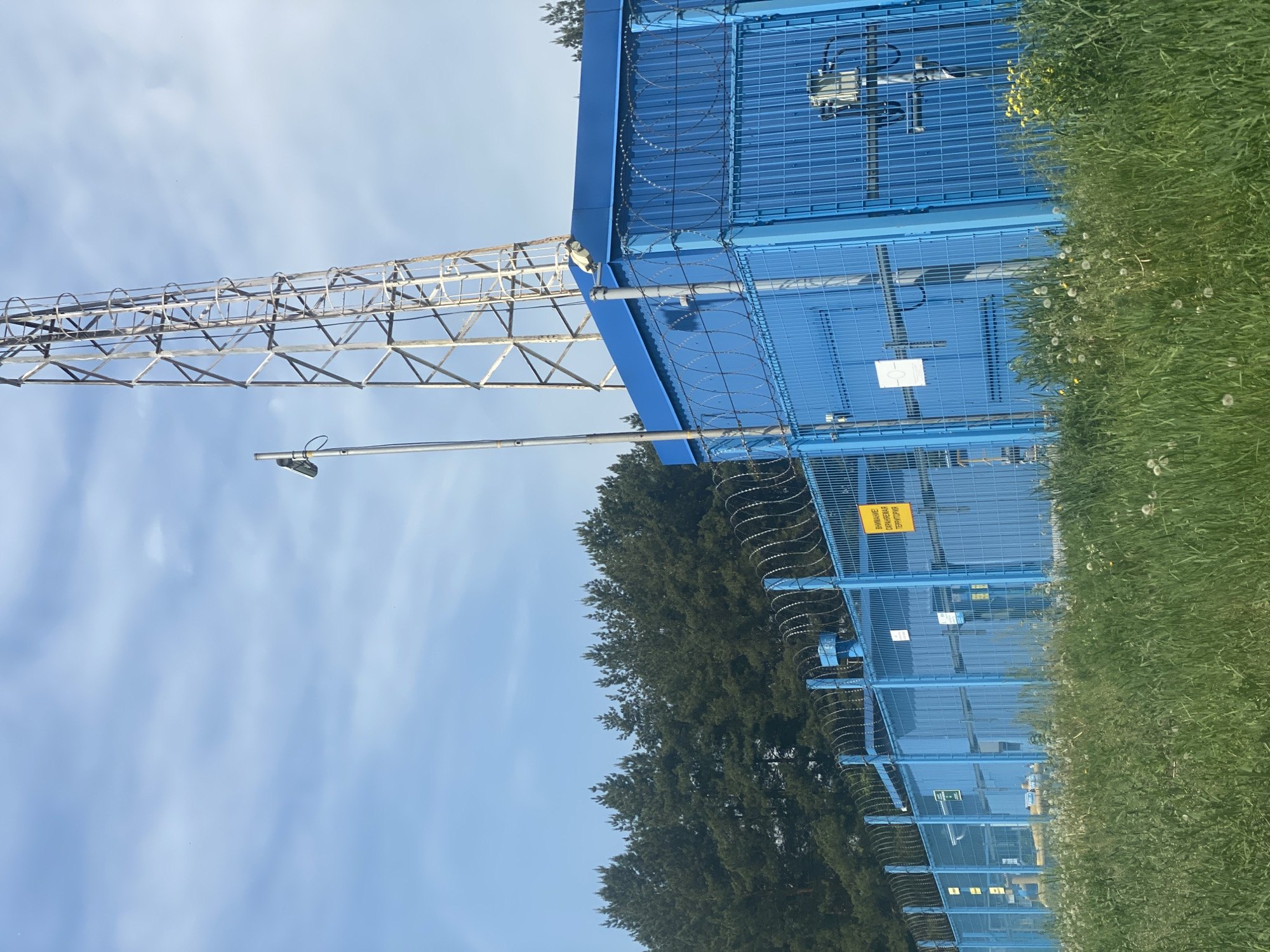 The image displays an outdoor scene in Novokuznetsk, Russia, under a light blue sky with scattered white clouds. The foreground consists of overgrown green grass interspersed with white dandelion seed heads and some yellow wildflowers.
Dominating the midground is a secure facility enclosed by a bright blue metal mesh fence, approximately 2-3 meters in height, topped with multiple strands of razor wire (concertina wire). A prominent yellow rectangular sign, written in Cyrillic, is affixed to the fence and reads: "ВНИМАНИЕ! ОХРАНЯЕМАЯ ТЕРРИТОРИЯ" (ATTENTION! PROTECTED AREA). Several smaller, white signs are also visible on the fence panels and interior structures. Behind the fence are multiple large, cuboid, blue modular structures or equipment enclosures. On the left side, behind the fence, sections of yellow industrial piping are visible. Various grey electrical boxes, cables, and sensor units are mounted on both the fence and the blue structures.
Adjacent to the blue facility, a tall, slender metal pole equipped with a security light or camera extends upwards. To the right of this pole, a tall, weathered metal lattice tower, likely a communication or transmission tower, rises into the sky, featuring a caged access ladder along one of its sides.
The background is characterized by a dense treeline of dark green pine trees, situated behind the secure facility. No people or animals are present in the image.