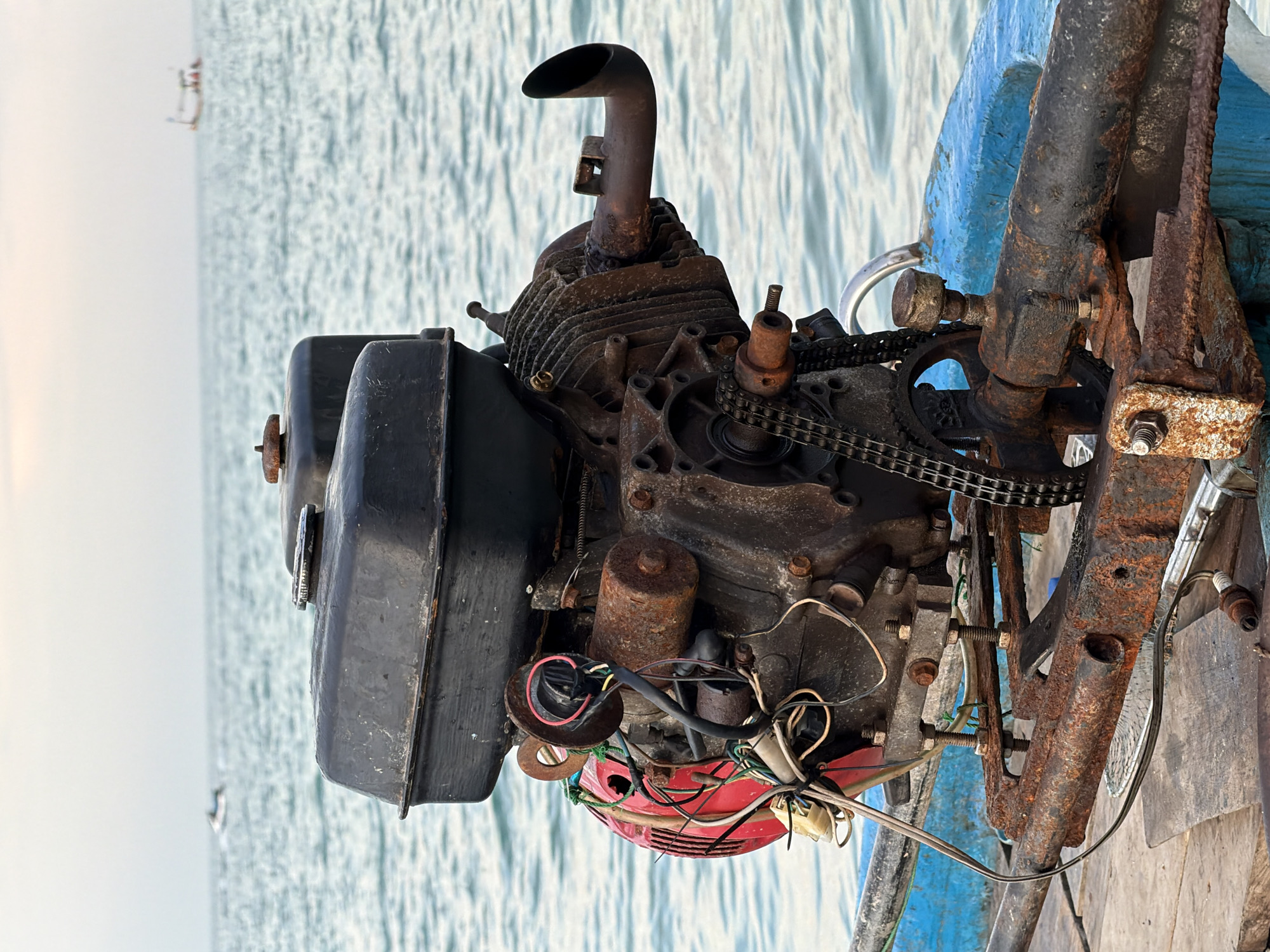 A rusted, vintage outboard motor sits on the deck of a small wooden boat. The motor, black with some red components visible, is heavily corroded and appears to be disused. The boat is on the water, with a hazy sky and the ocean stretching to the horizon. In the distance, a jet ski can be seen on the left, and a small fishing boat is visible on the right. The scene is likely located in the coastal waters of Ko Samui District, Thailand, given the tropical marine environment and the type of boat and motor.