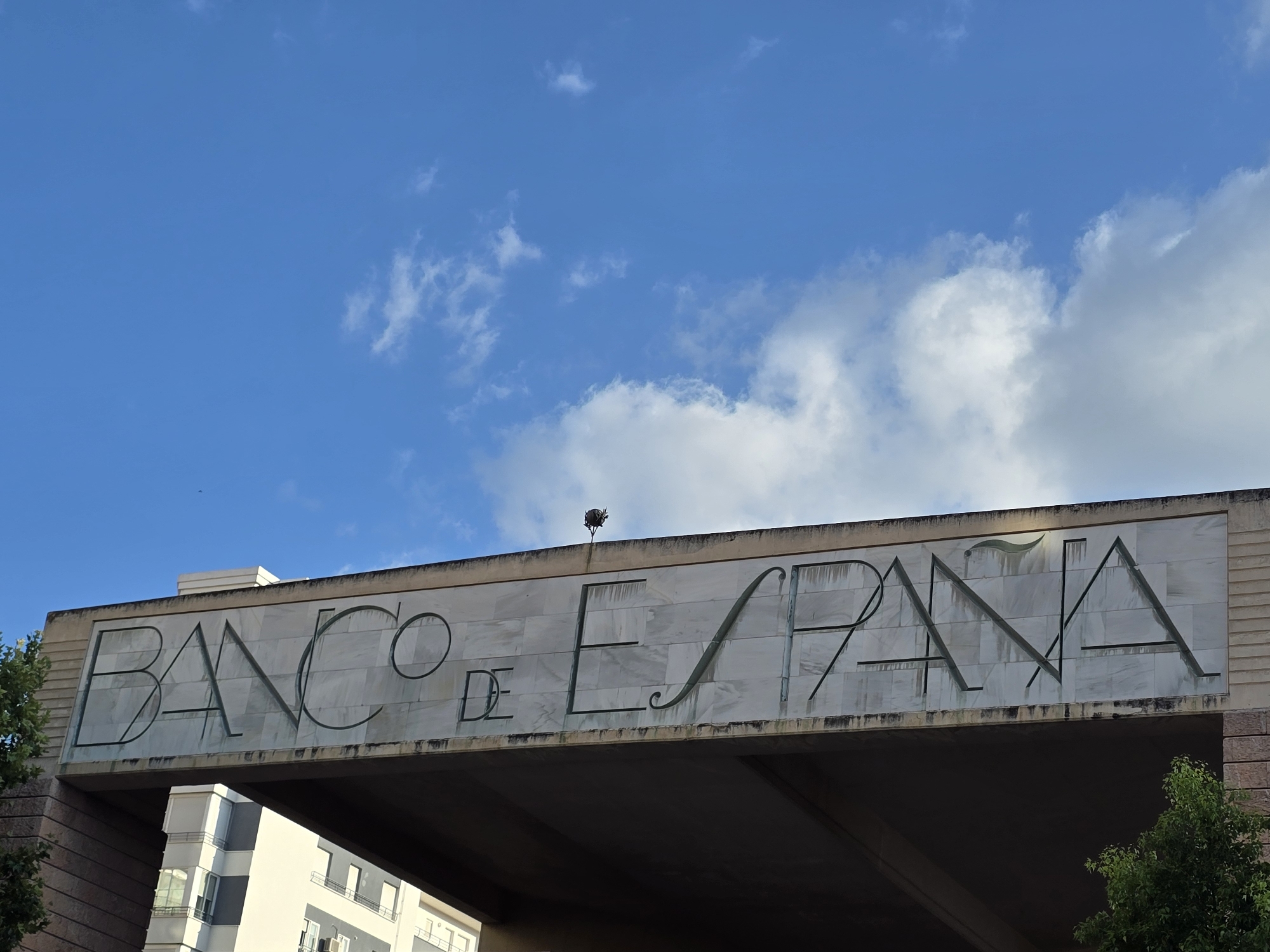 The image shows the facade of the Banco de España building in Jaén, Spain. The building features a large, marble-clad horizontal structure with the words "BANCO DE ESPAÑA" inscribed in large, stylized letters. The sky above is blue with scattered white clouds. A small dark object, possibly a bird or a decorative element, is perched on the top edge of the inscribed sign. In the lower part of the image, the dark underside of the overhanging structure is visible, and part of a modern white and grey building is seen to the left, with trees to the left and right.
