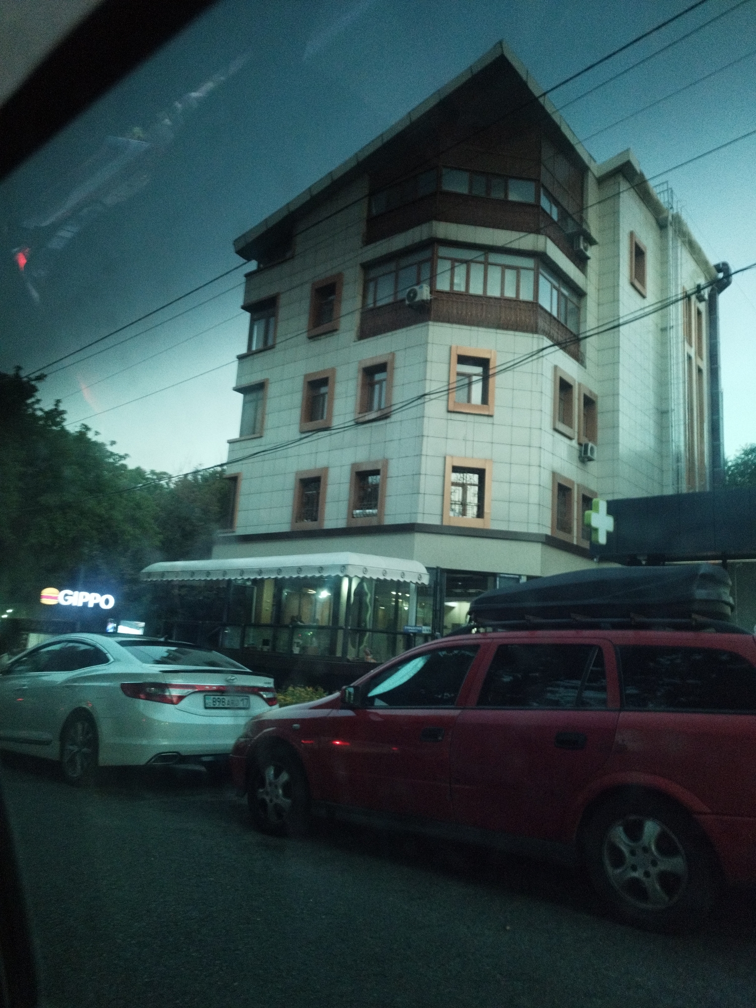 The image depicts a street scene in Shymkent, Kazakhstan. In the foreground, a red station wagon with a roof-mounted cargo box is parked on the right. To its left, a white sedan is also parked. Behind the vehicles, a multi-story building with a modern architectural design stands. The building has a beige facade with brown accents around the windows and along the roofline. A white awning extends from the building over a sidewalk area. A sign with the text "GIPPO" is visible to the left of the awning. A green cross symbol is mounted on a dark rectangular structure to the right of the awning. The sky is a deep blue with some faint clouds, suggesting it might be evening or a cloudy day. The overall impression is an urban environment with commercial establishments and residential areas.