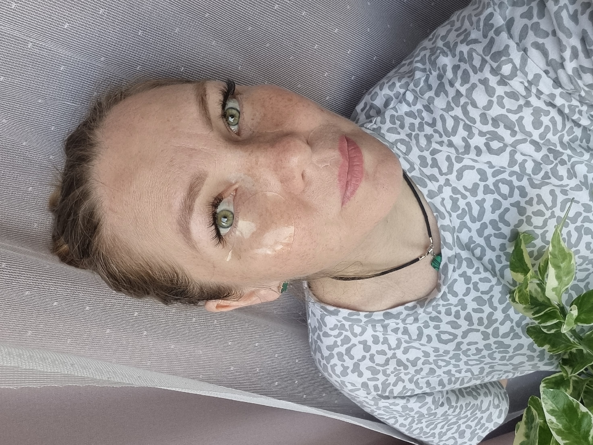 The image shows a close-up, waist-up, selfie-style portrait of a woman with fair skin, freckles, and green eyes. She has her hair pulled up in a bun and is wearing a light-colored t-shirt with a grey leopard print. She has on eye patches under her eyes and a dark necklace with a green pendant. She is looking slightly to her right. The background consists of a sheer, textured grey curtain. The image is an indoor photograph. There is no discernible information to associate this image with the city of Chaikovsky, Russia.