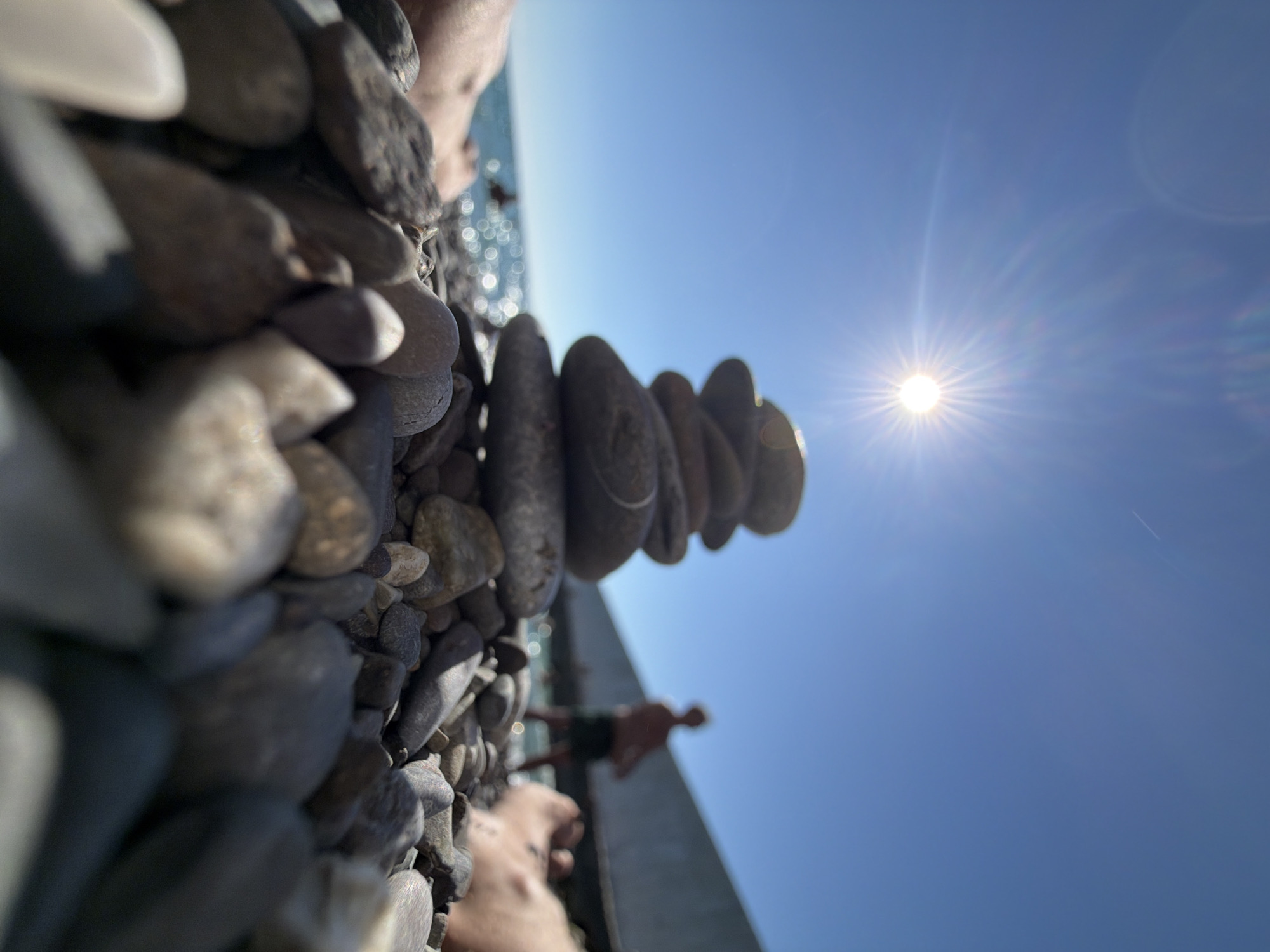 A low-angle shot captures a stack of smooth, gray stones balanced on a pebble beach under a bright, sunny sky. The sun, a radiant starburst, dominates the upper portion of the frame. In the background, the turquoise water of the sea sparkles, and a blurred figure of a person stands near a concrete structure. A hint of another person's leg is visible in the lower left corner. The overall scene suggests a peaceful day at the beach, likely in a coastal area such as Sochi, Russia, given the commonality of pebble beaches and coastal structures there. The focus is sharpest on the stones in the foreground, with the background elements progressively out of focus.