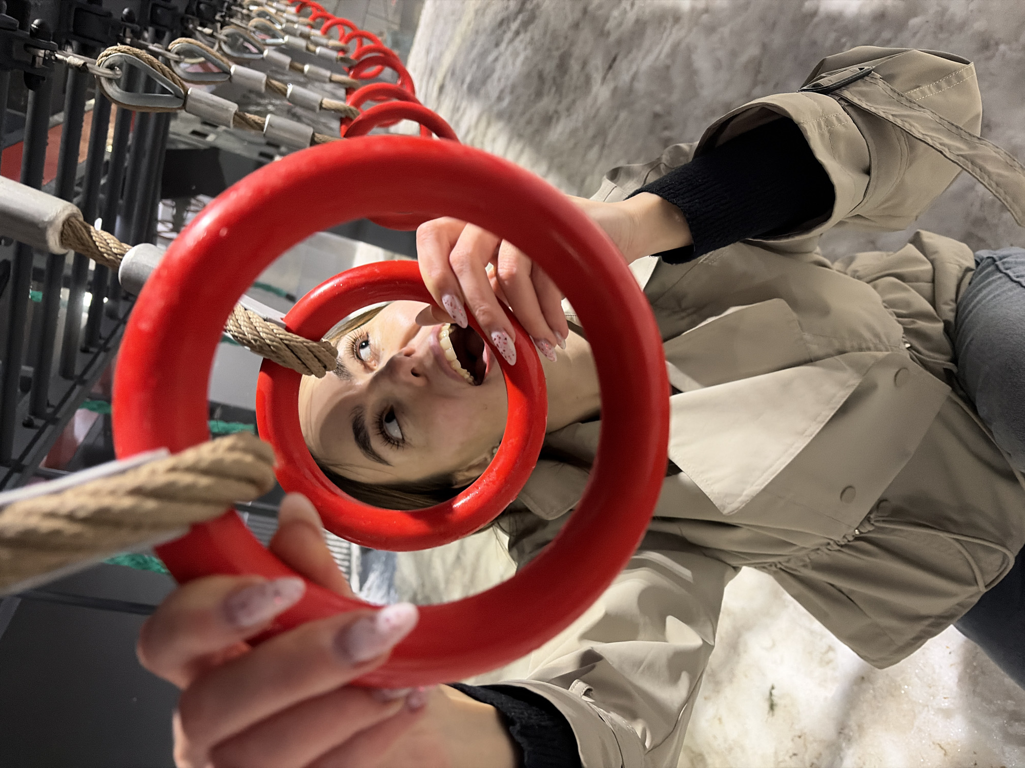 The image shows a young woman holding a red gymnastic ring in front of her face, with a row of similar rings stretching into the background. The rings are suspended by ropes attached to a metal structure. The woman has fair skin, light brown hair, and painted nails. She is wearing a beige trench coat over a black top and blue jeans. Her mouth is open, and she is looking upwards towards the right. The background includes snow on the ground. Based on the location and the nature of the equipment, it is likely the photo was taken at the 'Park of Culture and Rest named after S.M. Kirov' park in Izhevsk, Russia.