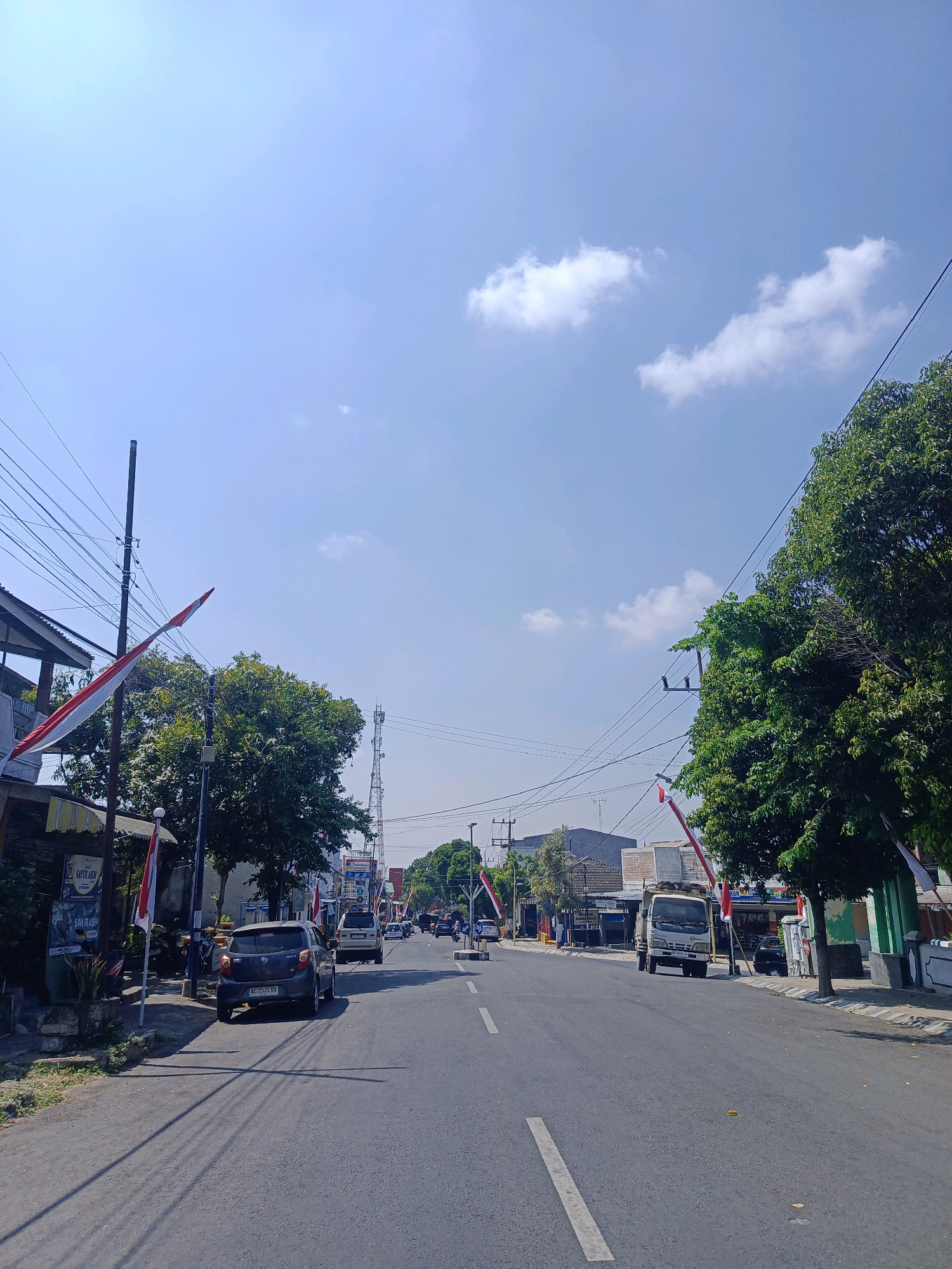 The image depicts a street scene in Kepolorejo, Indonesia, under a bright, clear blue sky with scattered white clouds. The road is paved and has a white line marking the center. On the left side of the road, there are buildings with awnings, trees, and Indonesian flags displayed. Several cars are parked along the side of the road.
Further down the street, there are more buildings, including what appears to be shops and a cell tower in the distance. Traffic is light, with a few cars and a small truck visible on the road. Trees line both sides of the street, providing shade. The overall impression is of a typical street in a developed area of Indonesia on a sunny day.