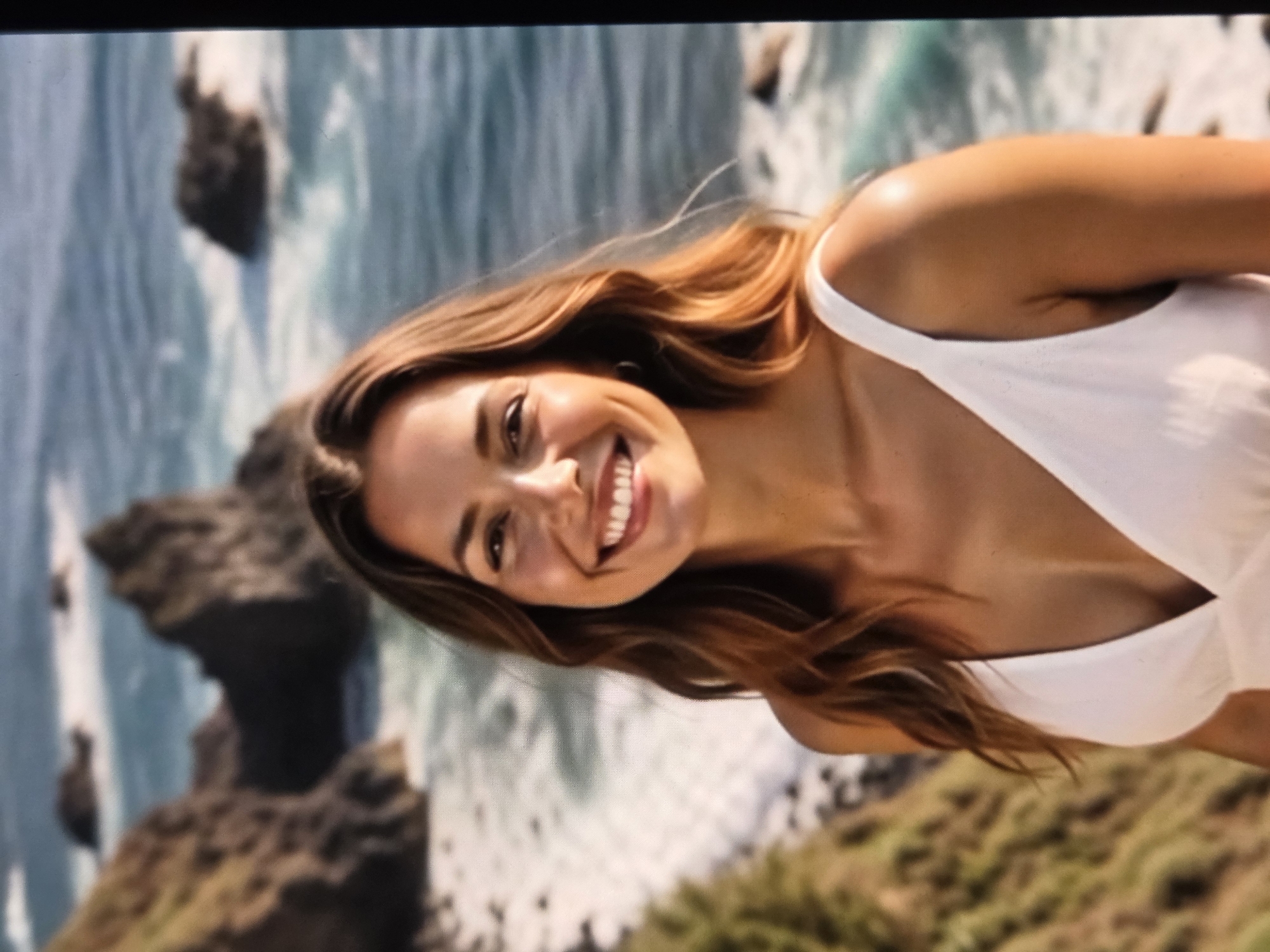 The image depicts a smiling woman with auburn hair wearing a white top. The background shows a coastline with rocks and ocean, as well as green vegetation. The location cannot be determined as Sofia, Bulgaria.