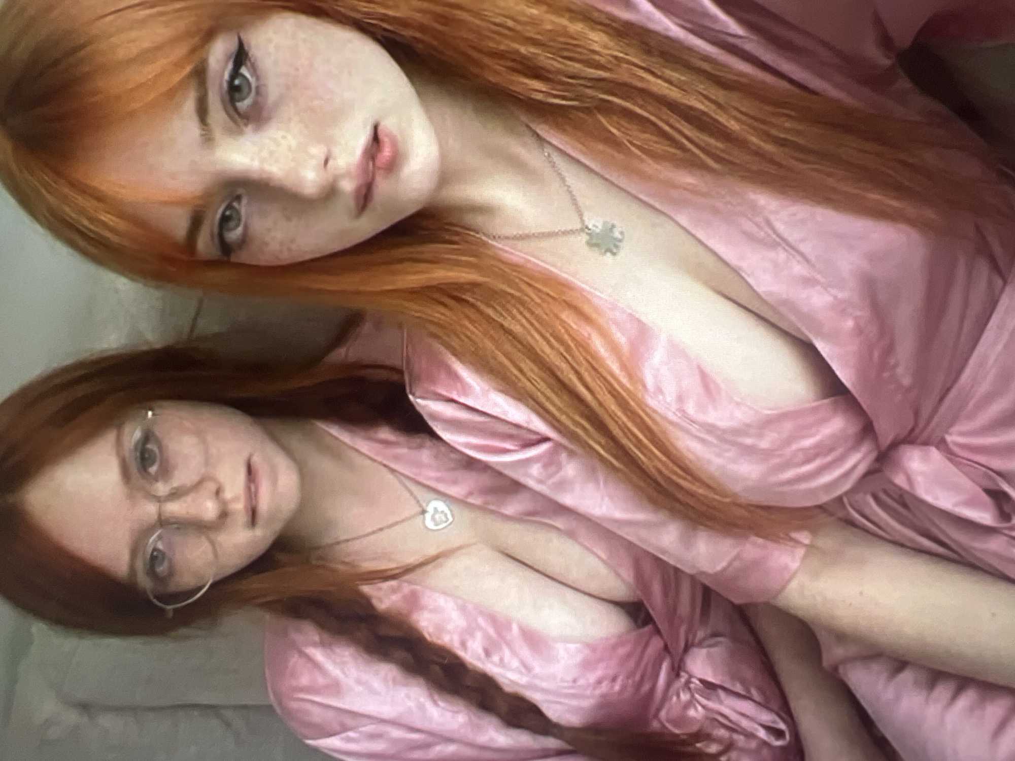 Two young women with long, reddish-orange hair are posing for a selfie. Both are wearing pink satin robes, with the woman on the right wearing a necklace with a clover-shaped pendant, and the woman on the left wearing a heart-shaped pendant necklace. The woman on the left is wearing glasses. They are positioned close together, with the woman on the right slightly in front of the woman on the left. The background is out of focus and appears to be an indoor setting, possibly a bedroom. There is no indication of the location being the city of Minsk, Belarus.