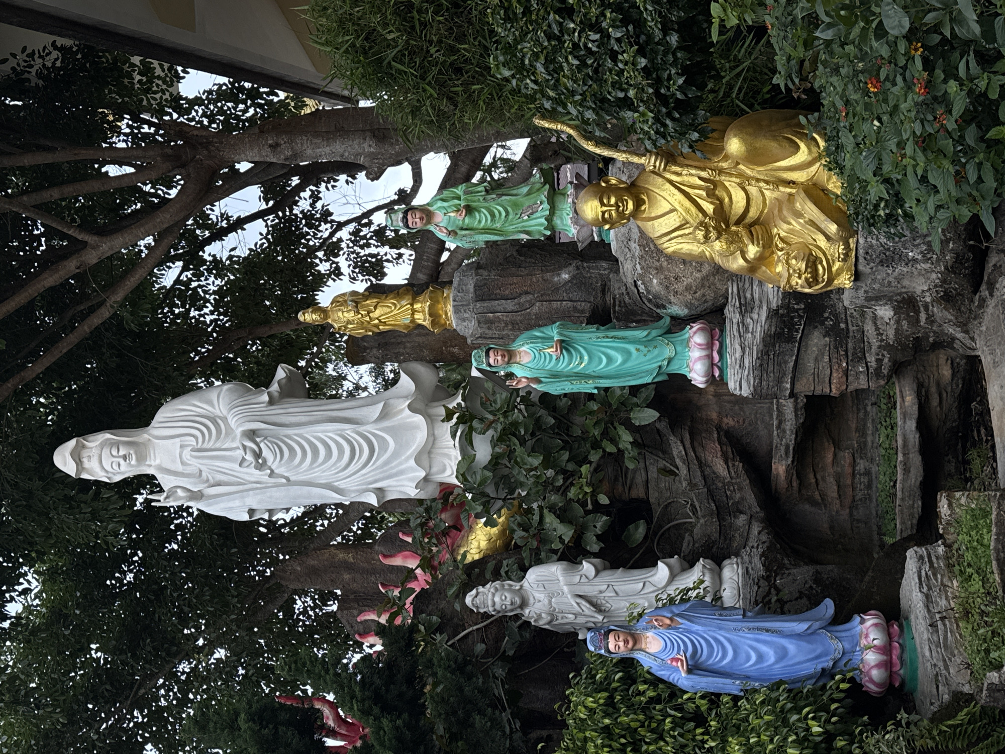 The image depicts a series of Buddhist statues in Lâm Viên - Đà Lạt, Vietnam. The statues, arranged vertically, are diverse in color and style. From top to bottom, there is a golden statue, a green statue, a small golden statue, a light-green statue, a tall white statue, a golden statue, a light-gray statue, and a blue statue resting on a lotus flower. The statues are surrounded by lush greenery, trees, and vegetation, indicating an outdoor setting within a garden or park. The background features branches of a large tree reaching towards the sky.