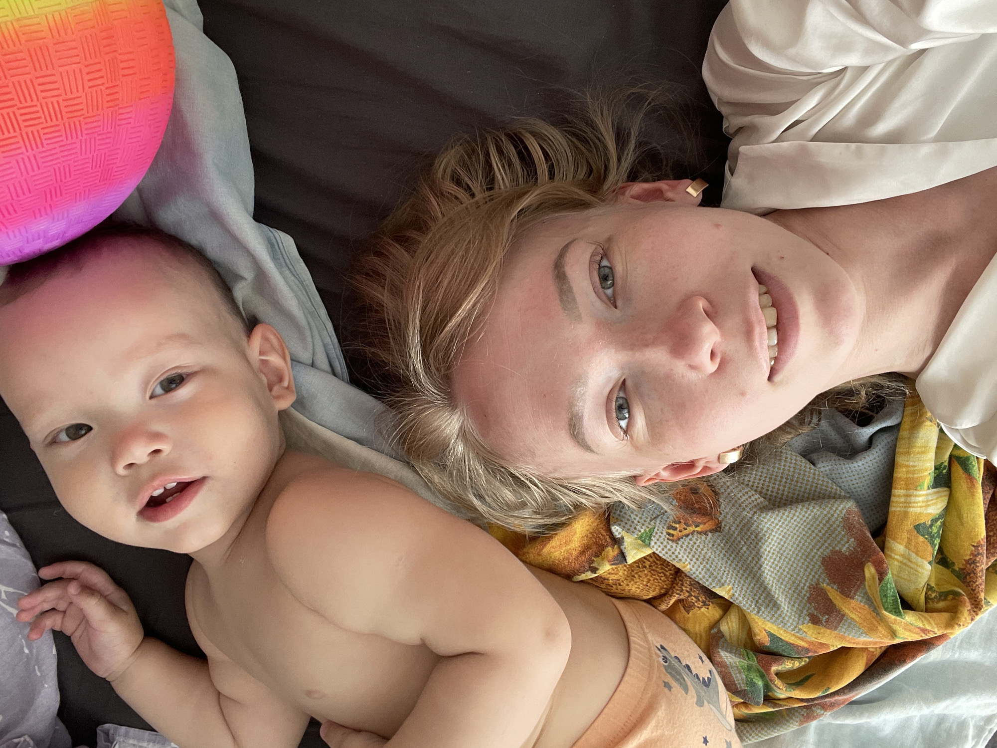 A woman and a baby are lying on a bed, potentially indoors. The woman has fair skin, blonde hair, blue eyes, and is wearing a gold earring. She is smiling slightly. The baby has fair skin, light brown hair, and is wearing an orange diaper with a small dinosaur print. A multicolored ball rests near the baby's head. The bed has various fabrics, including a yellow-and-brown patterned scarf, a light blue sheet, and a dark gray sheet. There is no indication of the location being Perm, Russia, based on visual cues.