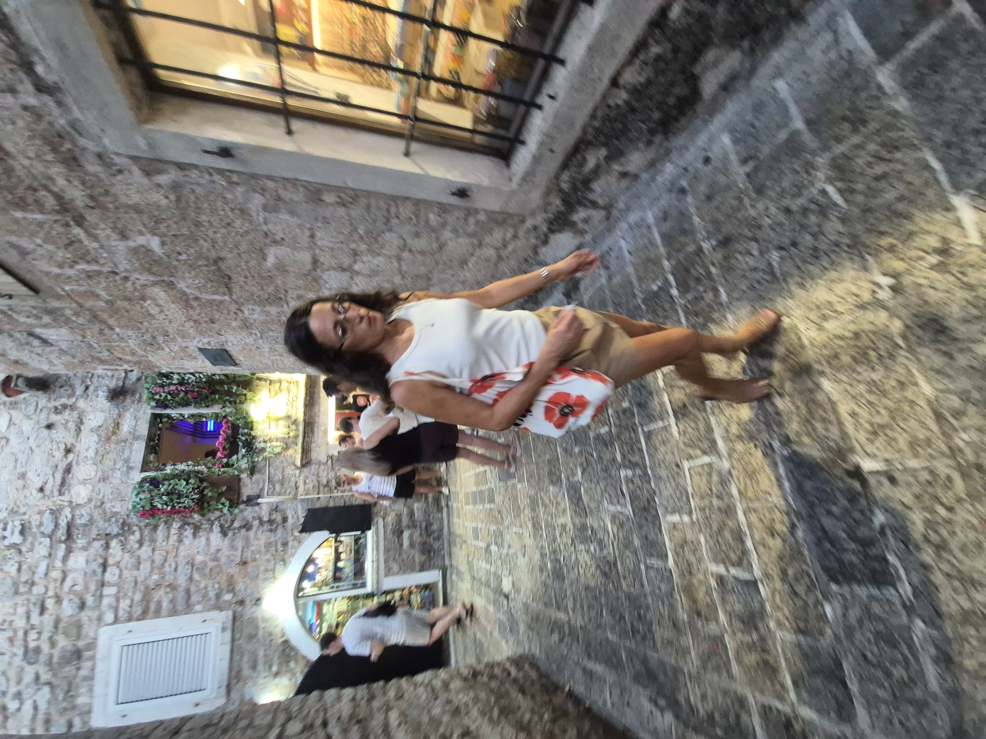 The image depicts a woman walking down a narrow, cobblestone street in the Old Town of Budva, Montenegro. She is wearing a white tank top, a tan skirt, and sandals, and carrying a white tote bag with red poppy designs. The street is lined with stone buildings, some with flower boxes on the windows. In the background, other people can be seen walking and browsing in shops. The lighting suggests it is either early morning or late evening. The scene captures a typical street view within the historic part of Budva.