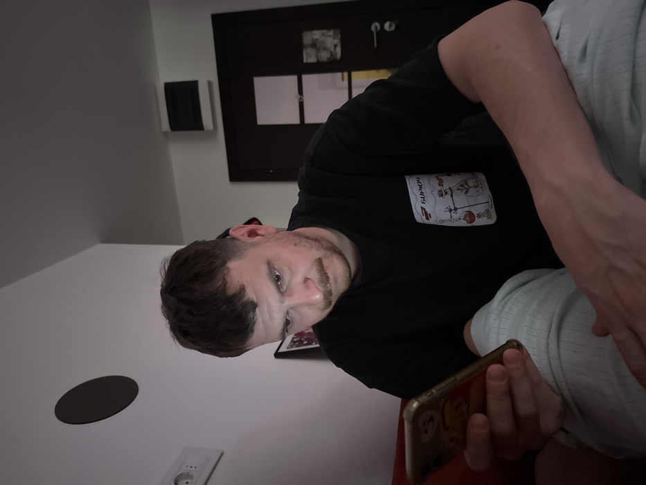 A man with short brown hair and a stubbly beard reclines on a bed, holding a gold smartphone. He wears a black t-shirt with a white graphic patch on the chest. Behind him is a dark-colored door and a small black square mounted on the white wall. The room appears to be a hotel room. A round brown object and a power outlet are visible on the lower-left side of the frame. The photo is shot from a slightly elevated angle, with a tilted orientation.