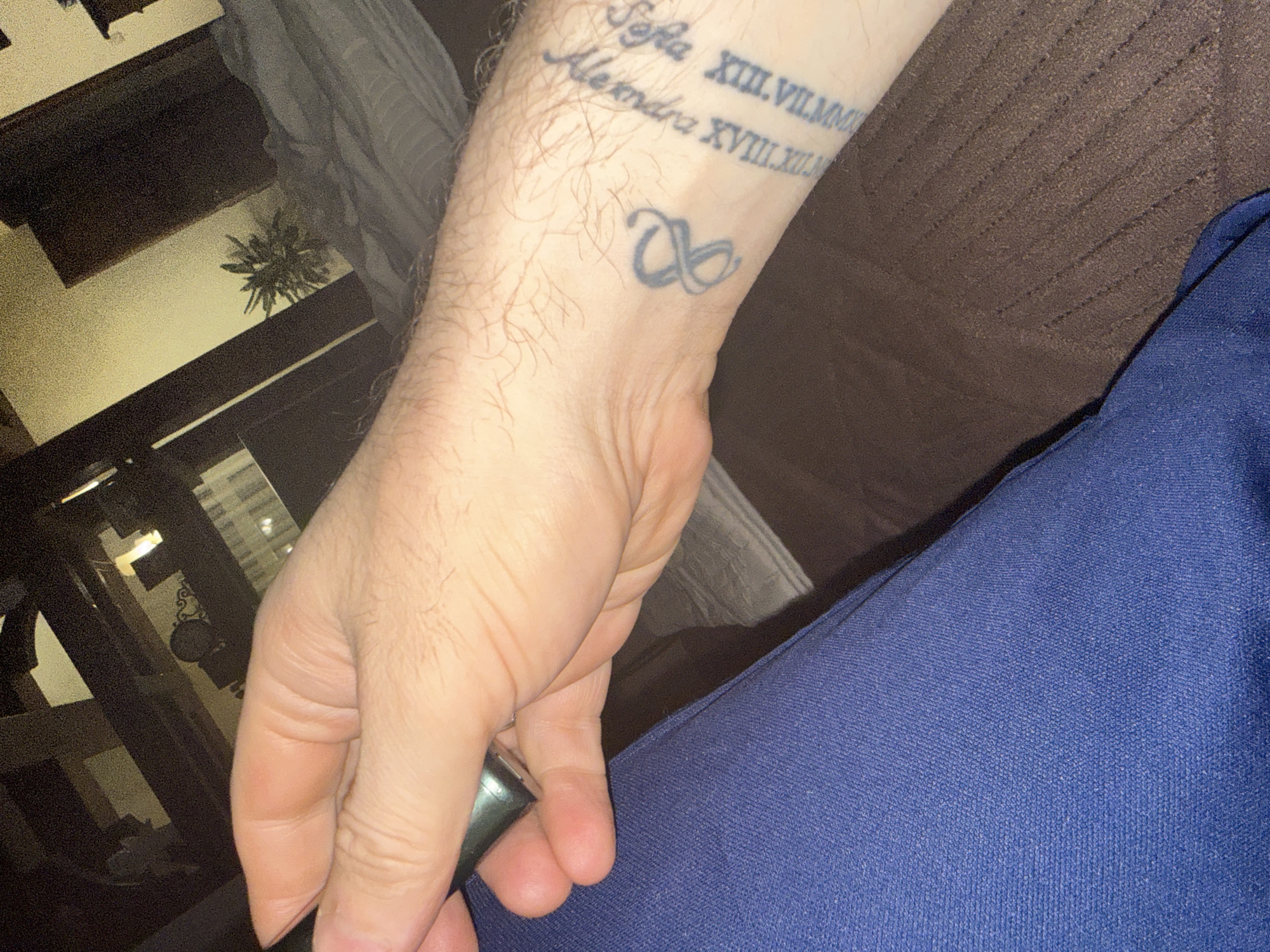 A close-up photograph depicts a Caucasian male's left forearm and hand. The forearm bears two tattoos: one displaying the names "Sofia" and "Alexandra" accompanied by Roman numerals (XIII.VII.MM and XVIII.XIII.XII respectively), and another a looped design near the wrist. The hand is pale and lightly haired, holding an indiscernible metallic object. The subject is wearing blue clothing. The background includes a textured brown sofa, a glimpse of a wooden furniture item, and a dark shadow, possibly indoors in Lesnoi Gorodok, Russia.