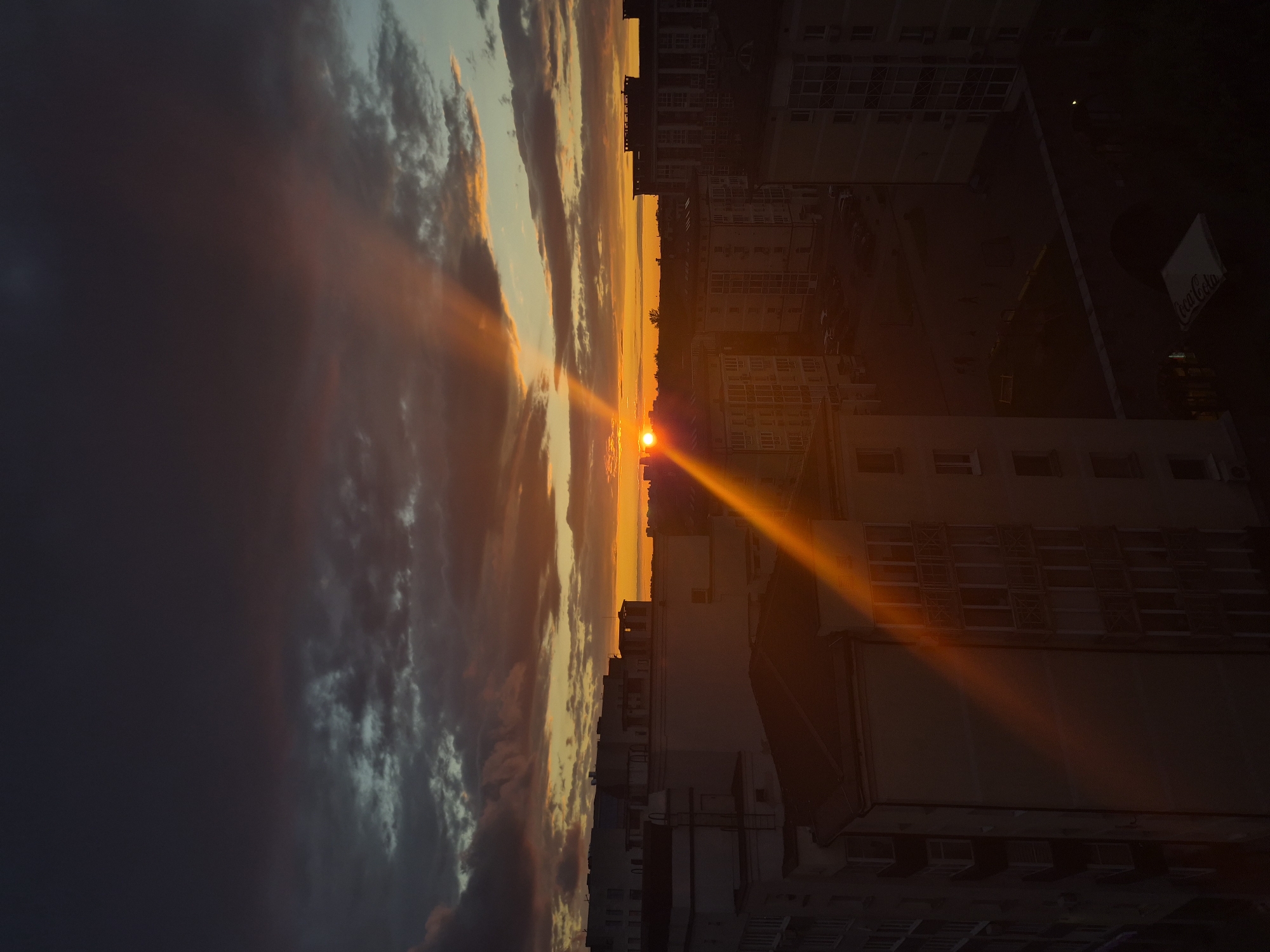 An aerial view of a city skyline at sunset, with dramatic rays of sunlight piercing through dark clouds. The sun, a bright orange orb, is low on the horizon, casting long shadows and illuminating the sky with vibrant hues of orange and yellow. The foreground is dominated by apartment buildings of varying heights, their facades partially silhouetted against the colorful sky. Some buildings have balconies and windows visible, suggesting residential occupancy. In the distance, more buildings stretch towards the horizon, creating a dense urban landscape. The street level is barely discernible, with hints of parked cars and possibly some pedestrians. The overall impression is one of a serene, yet dynamic, urban environment bathed in the warm glow of a setting sun. The scene appears to be of Minsk, Belarus, given the architectural style and the characteristic sky formations often seen in the region.