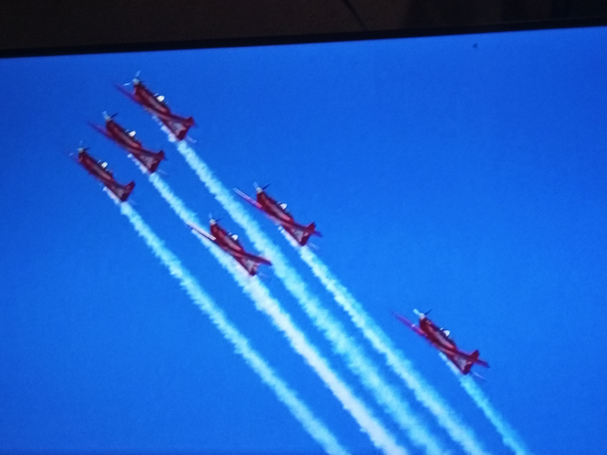 Six red airplanes in formation, trailing white smoke against a bright blue sky, are captured in this aerial photograph. The planes are arranged in a diagonal line, moving from the upper left to the lower right of the frame. The perspective suggests the image was taken from a higher vantage point, possibly from another aircraft or drone. The scene is devoid of any ground features, focusing solely on the aircraft and their contrails. There is no visible indication or context within the image to suggest a specific location in Banyudono, Indonesia.