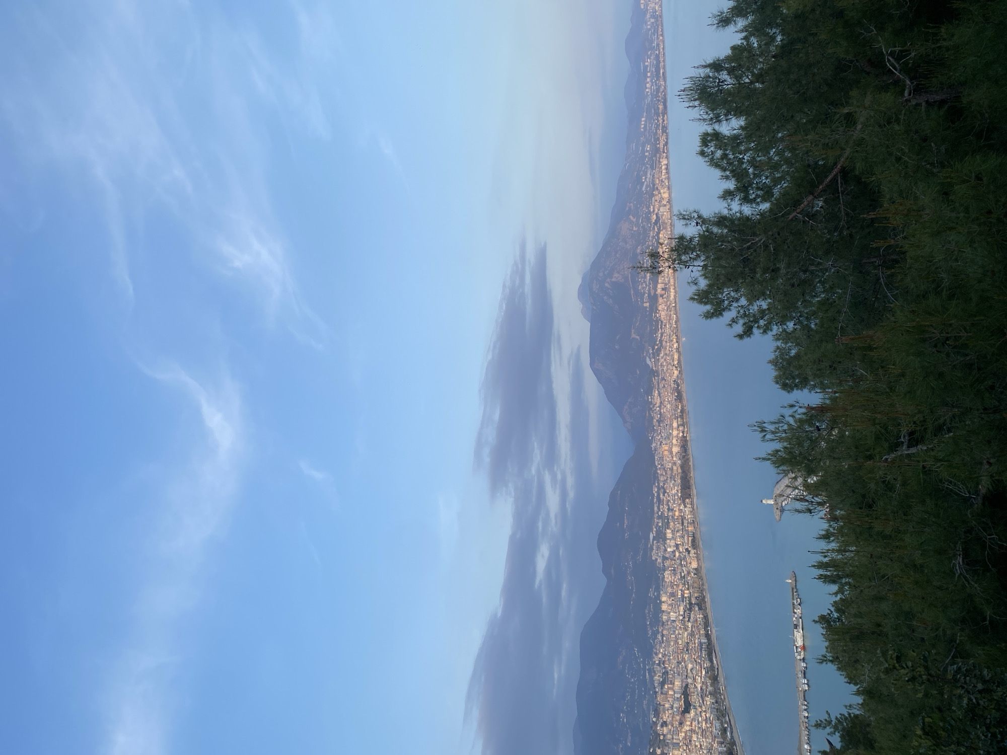 A panoramic view from an elevated position in Alanya, Turkey, depicts a long, coastal city situated between the sea and mountainous terrain. The city is densely built, with buildings closely aligned along the shoreline. Mountains rise behind the city. Clouds are visible over the mountains and in the sky. In the foreground, the edge of a forested area is partially visible. A port area with cranes and ships is visible in the water. The sky is a clear blue with wisps of clouds.
