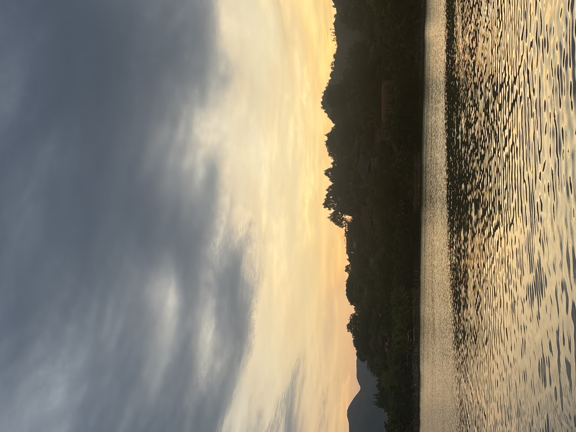 A serene lake is in the foreground, reflecting the warm glow of the setting sun. The water is calm with gentle ripples, creating a mesmerizing pattern of light and shadow. In the background, a lush green hillside is silhouetted against the vibrant sky, dotted with trees and a few scattered buildings. The sky itself is a gradient of soft oranges and yellows near the horizon, transitioning to a deep blue with streaks of gray clouds higher up. The overall atmosphere is peaceful and tranquil, capturing the beauty of nature at dusk. The image does not contain any people or specific identifiable landmarks.