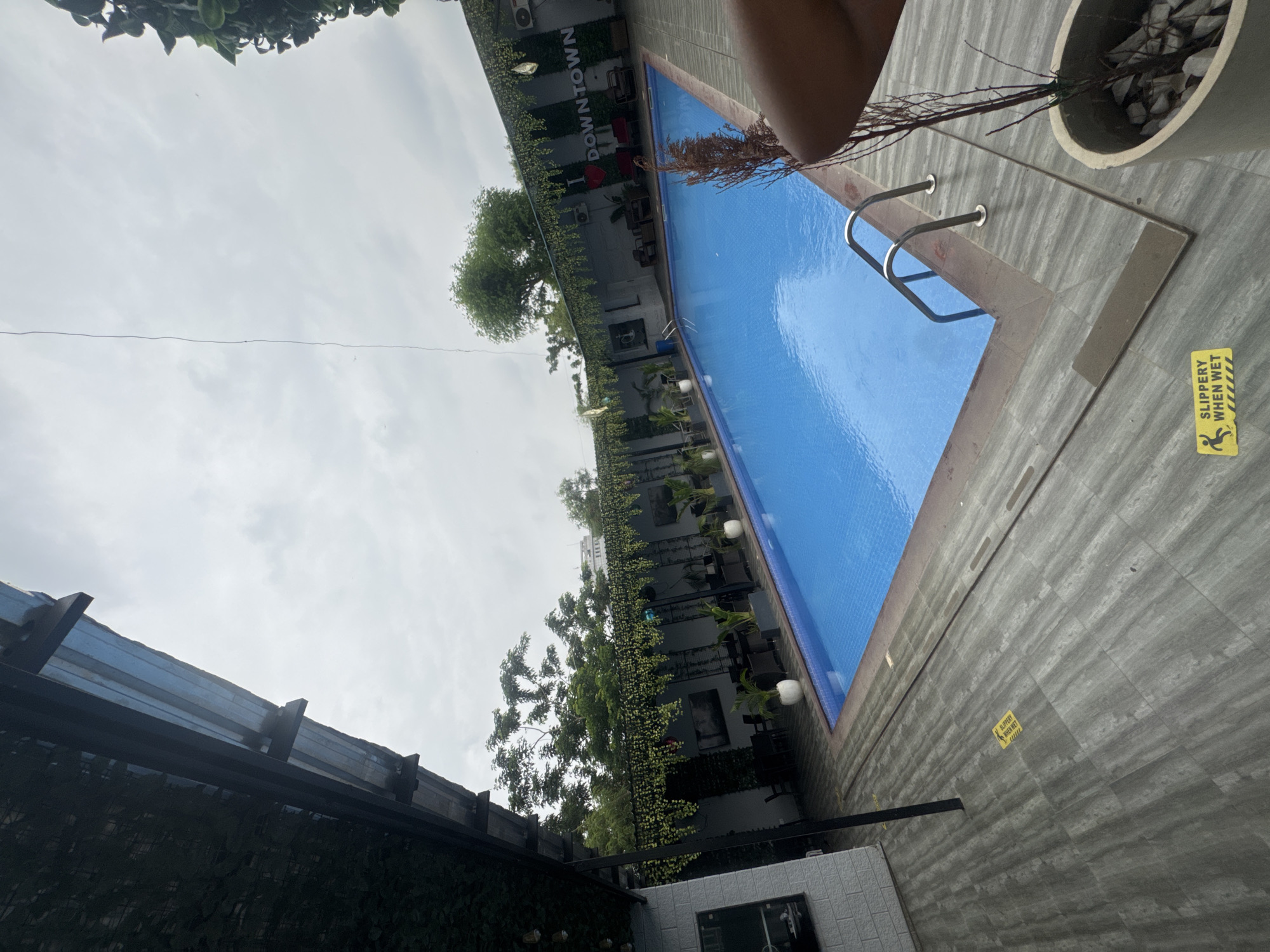 The image shows a swimming pool area in what appears to be an urban setting. "I <3 DOWNTOWN" is written on the wall. The pool is rectangular and filled with blue water, and has a metal ladder at one end. Adjacent to the pool is a tiled patio with a "Slippery When Wet" sign. Potted plants are scattered around the pool area. In the background, there's a building with greenery growing on its walls. The sky is overcast. No people are clearly visible in the main scene, except for parts of a person at the top of the image.