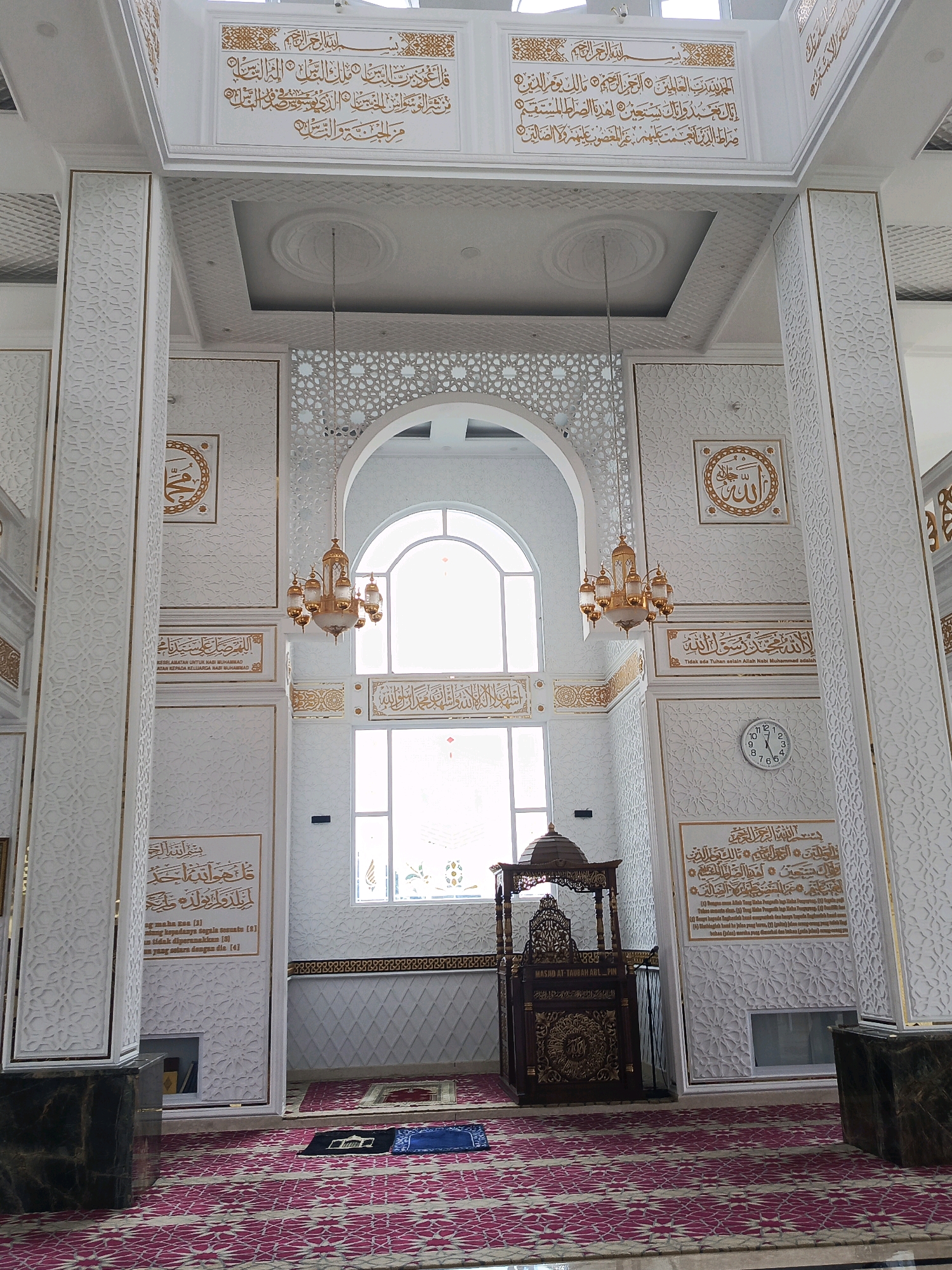 The image shows the interior of a mosque in Cisarua, Indonesia, characterized by its white walls adorned with gold Arabic calligraphy and intricate geometric patterns. The central focus is a large arched window, through which daylight streams into the prayer hall. Two ornate chandeliers hang from the ceiling, illuminating the space. In front of the window, a wooden minbar, or pulpit, with detailed carvings stands prominently. The floor is covered with a red and pink patterned carpet, and two prayer mats are laid out, suggesting recent or imminent prayer. Large white pillars with decorative gold accents support the high ceiling, which features recessed lighting and further ornate designs. The overall impression is one of grandeur and solemnity, typical of a well-maintained Islamic place of worship.