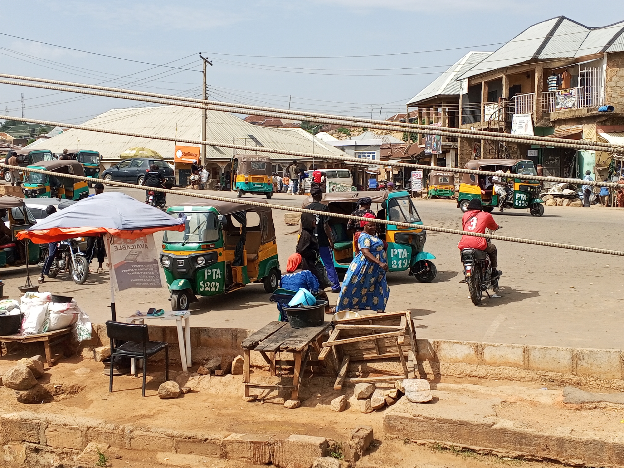 The image depicts a street scene in Gwong, Nigeria, characterized by a bustling commercial environment. Several green and yellow auto-rickshaws, identified by "PTA" markings and varying numerical identifiers (e.g., PTA 5227, PTA 2195, PTA 6550), are visible.
A woman in a blue patterned dress stands near a makeshift wooden table laden with a black bucket and other containers. Nearby, there is an area for mobile money transactions, evidenced by a table, chair, and advertising display with the text "ZEND WOWEA...POS."
Several people are seen walking, standing, or interacting in the street. A motorcyclist wearing a red shirt rides past. In the background are buildings, including a two-story structure with a balcony, and various shops and businesses. Overhead, a network of cables traverses the street.