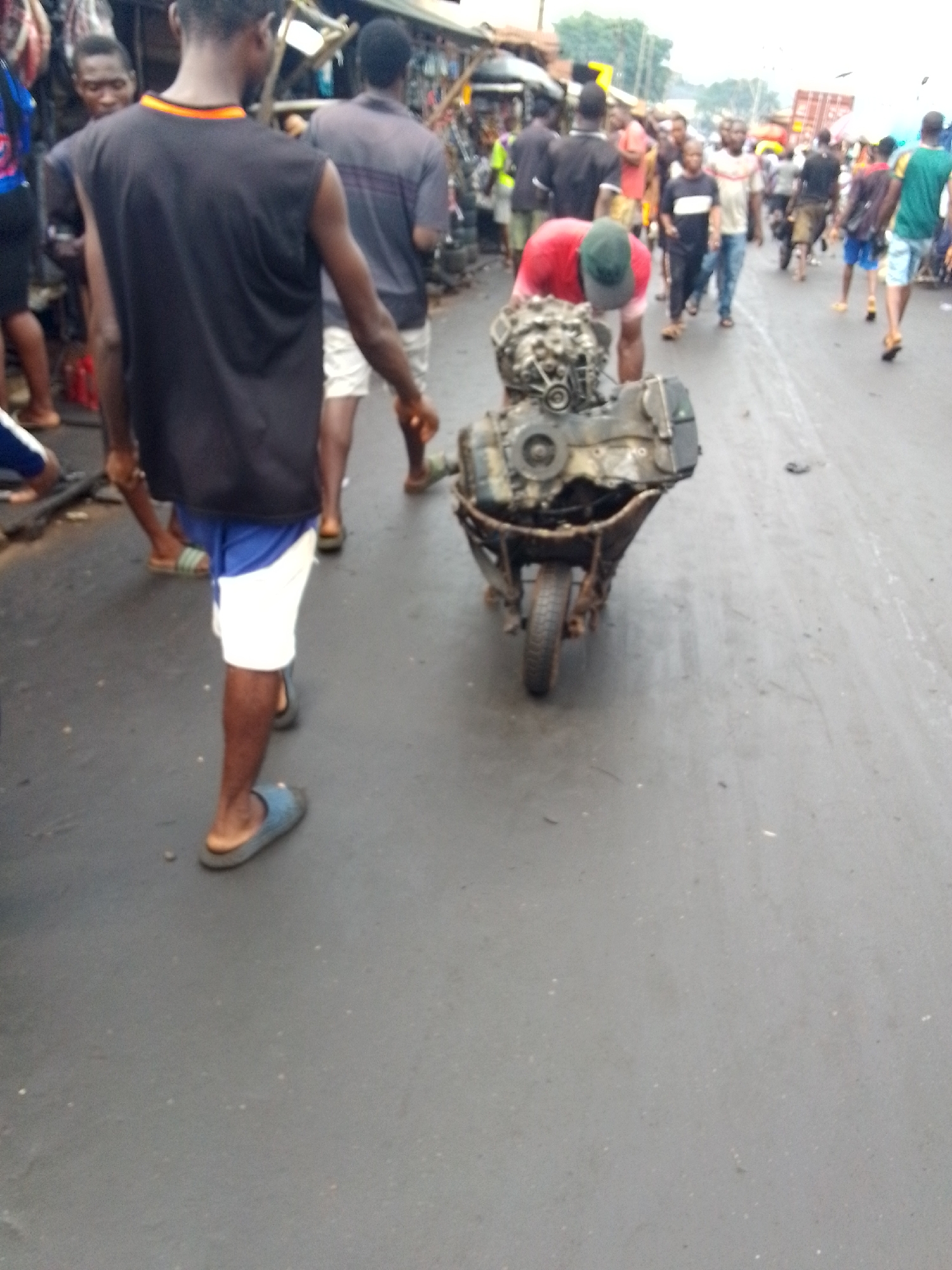 The image captures a bustling street scene in what appears to be a market or commercial area in Onitsha, Nigeria. The perspective is from behind a person walking, looking down the street.
In the immediate foreground, a man with dark skin, seen from the back, walks away from the viewer. He wears a black sleeveless shirt with an orange and yellow trim around the neck, and blue and white shorts. He has light blue slip-on sandals on his feet.
Slightly ahead and to the right, a man in a red shirt and a green baseball cap is bent over, pushing a rusty wheelbarrow. The wheelbarrow carries a large, greasy, metallic engine, possibly from a vehicle, indicating the transport of heavy machinery or parts.
The street itself is paved with dark grey asphalt, appearing somewhat damp or wet in sections. Numerous other people, mostly men, are visible in the mid-ground and background, walking in various directions, contributing to the busy atmosphere. Their attire is casual, including t-shirts, shorts, and trousers. On both sides of the street, there are stalls or informal structures, suggesting a marketplace where goods might be bought, sold, or repaired. In the far background, a large truck or container is partially visible, indicating vehicle traffic.
The lighting suggests it is daytime, though the sky appears overcast or the area is shaded, preventing harsh shadows. No specific emotions are discernible from the individuals. There is no visible text in the image.