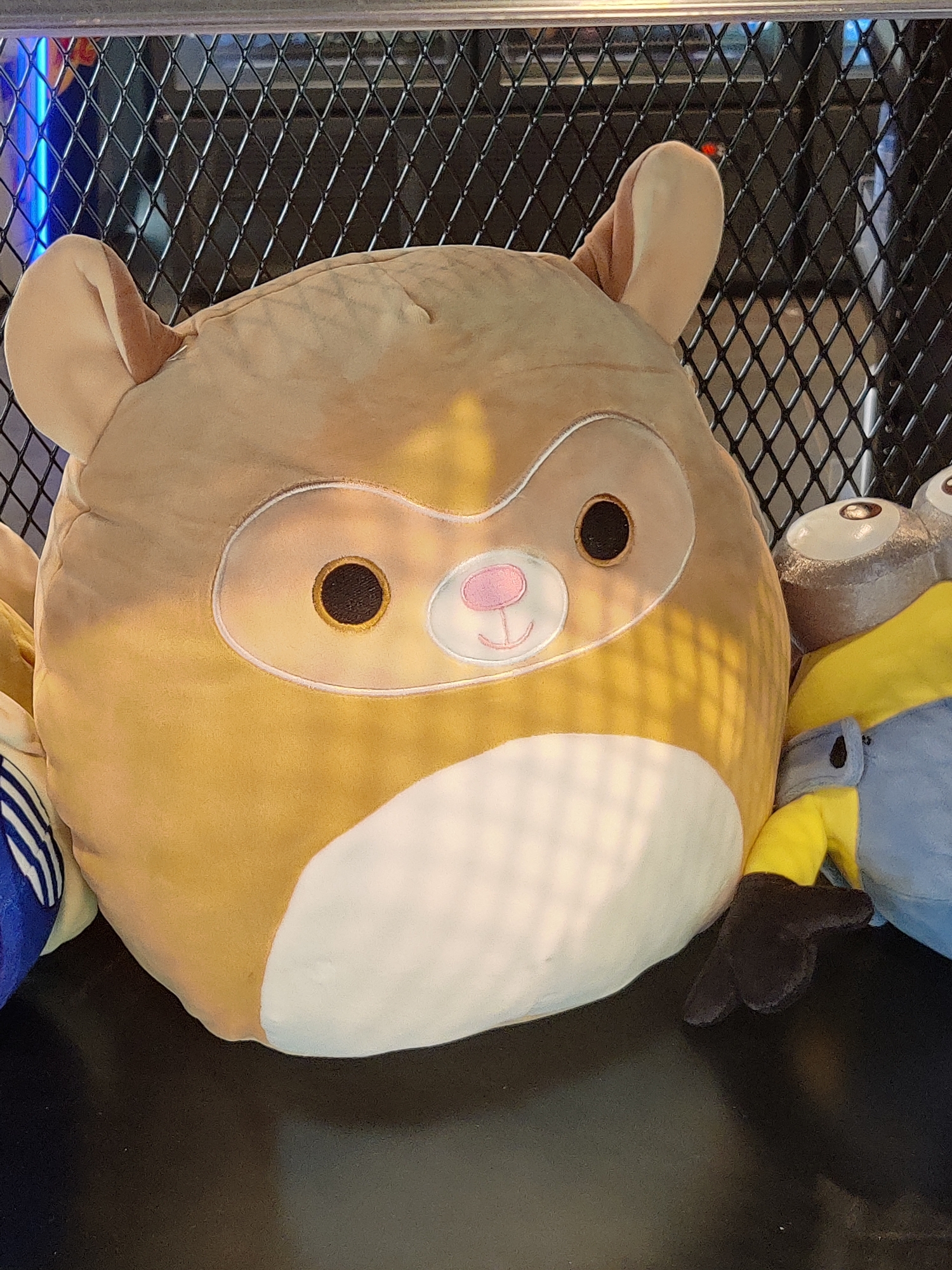 A close-up, eye-level, slightly angled shot shows several stuffed toys. A large, light brown stuffed animal with white accents on its face, resembling a sloth or monkey, is positioned in the foreground, slightly left of center. It has large, dark brown eyes, a pink nose, and two light brown ears. To the right of this animal, a portion of a Minion character from the "Despicable Me" franchise is visible, wearing blue overalls and a yellow shirt. The Minion has a silver-colored head and one visible eye. The stuffed toys are resting on a dark, smooth surface, possibly a table or shelf. Behind the toys, a black metal grid with diamond-shaped openings is present. In the upper left corner, a bright blue light emanates from behind the grid, casting a faint glow. The scene appears to be indoors, with artificial lighting and a dark background. There is no indication of the city of Eijsden, Netherlands, in the image itself.