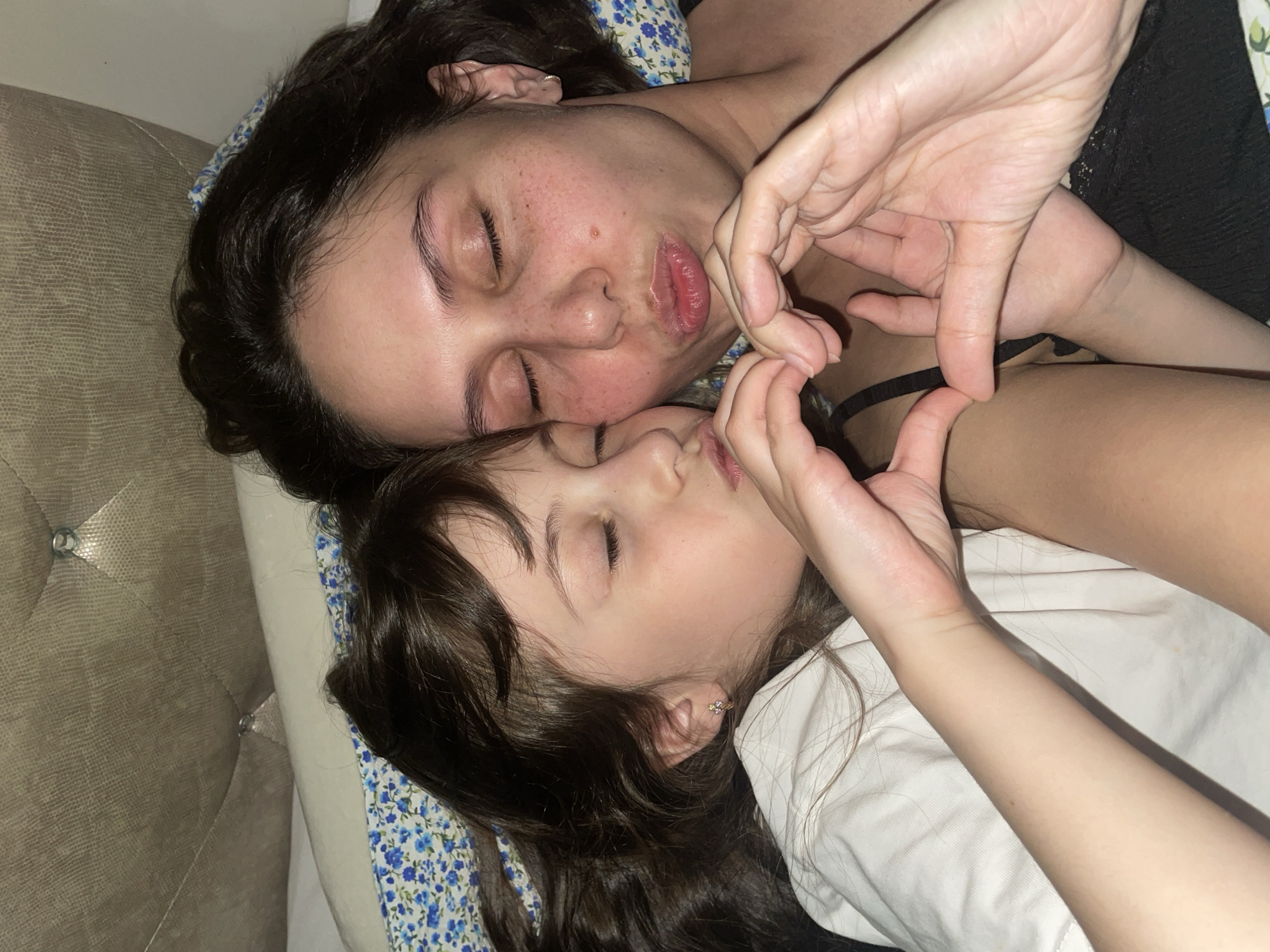 An overhead shot captures an adult and a child lying closely together, seemingly asleep or resting, on what appears to be a bed. The adult, with dark hair, is positioned on a pillow adorned with a blue floral pattern. Her eyes are closed, and her lips are slightly puckered in a gentle expression. Beside her, a child, also with dark hair, rests with closed eyes and a small earring in her ear.
A prominent feature is their intertwined hands, which are deliberately positioned to form a heart shape, symbolizing affection or love. The adult's hands form the top curves of the heart, while the child's hands complete the bottom. The adult is wearing a black-strapped garment, possibly a nightgown or top, while the child is dressed in a white shirt or covered by white bedding.
The setting is indoors, likely a bedroom, indicated by the floral pillow and a beige, upholstered headboard with crystal-like buttons visible on the left. The lighting is soft and dim, suggesting a peaceful moment, possibly at night or during a quiet downtime. The overall mood is one of intimacy, tenderness, and familial closeness. No text is visible in the image. The location context provided is Izhevsk, Russia.