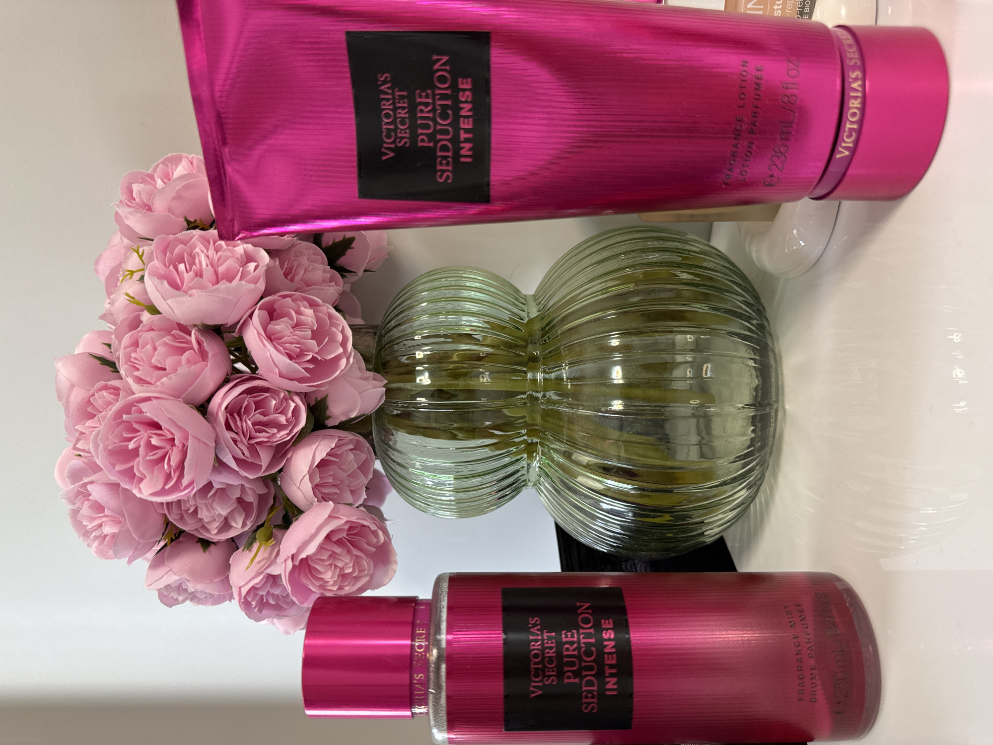 The image displays a tabletop scene featuring two Victoria's Secret Pure Seduction Intense products: a fragrance lotion in a tube and a fragrance mist in a bottle. The products are primarily pink with black labels. A glass vase with a ridged surface and filled with green liquid is centered between the beauty products and a bouquet of pink flowers to the left. The background is mostly white.