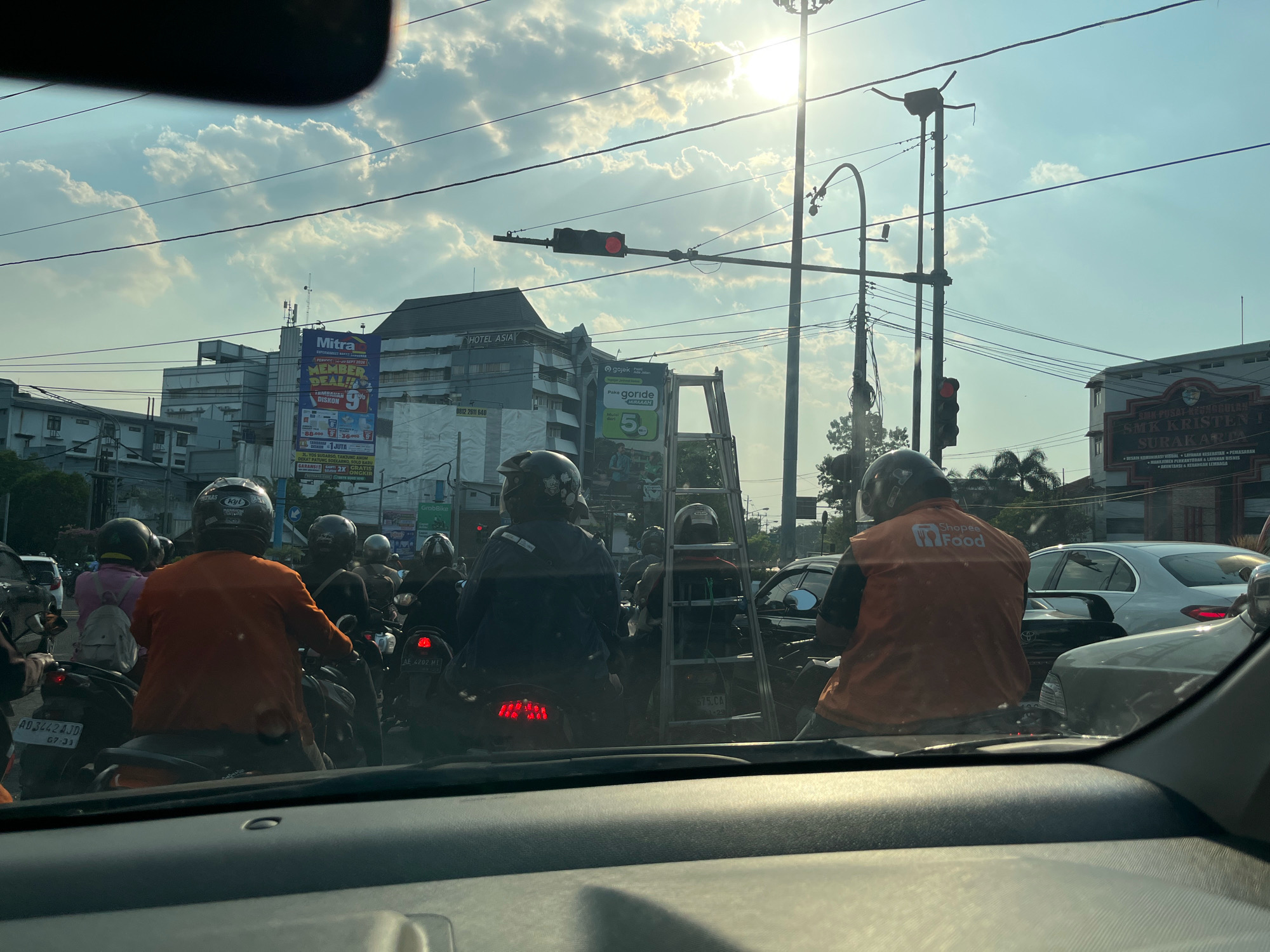 The image captures a traffic scene in the city of Surakarta, Indonesia, likely during daylight hours with the sun shining brightly, creating lens flare and casting shadows. Several motorcycles and cars are present, indicating a traffic jam or intersection. Many of the motorcyclists are wearing helmets and some are wearing orange vests with logos, suggesting they are delivery drivers for services like Shopee Food or Gojek. One individual in the foreground on a motorcycle is wearing an orange vest with "Shopee Food" printed on it. In the background, buildings are visible, including one labeled "HOTEL ASIA" and another with "SMK KRISTEN 1 SURAKARTA" signage. A tall ladder is strapped to the back of a motorcycle, possibly being transported by a worker. Traffic lights are visible, and one appears to be red. The overall impression is one of urban congestion and daily activity in Surakarta.