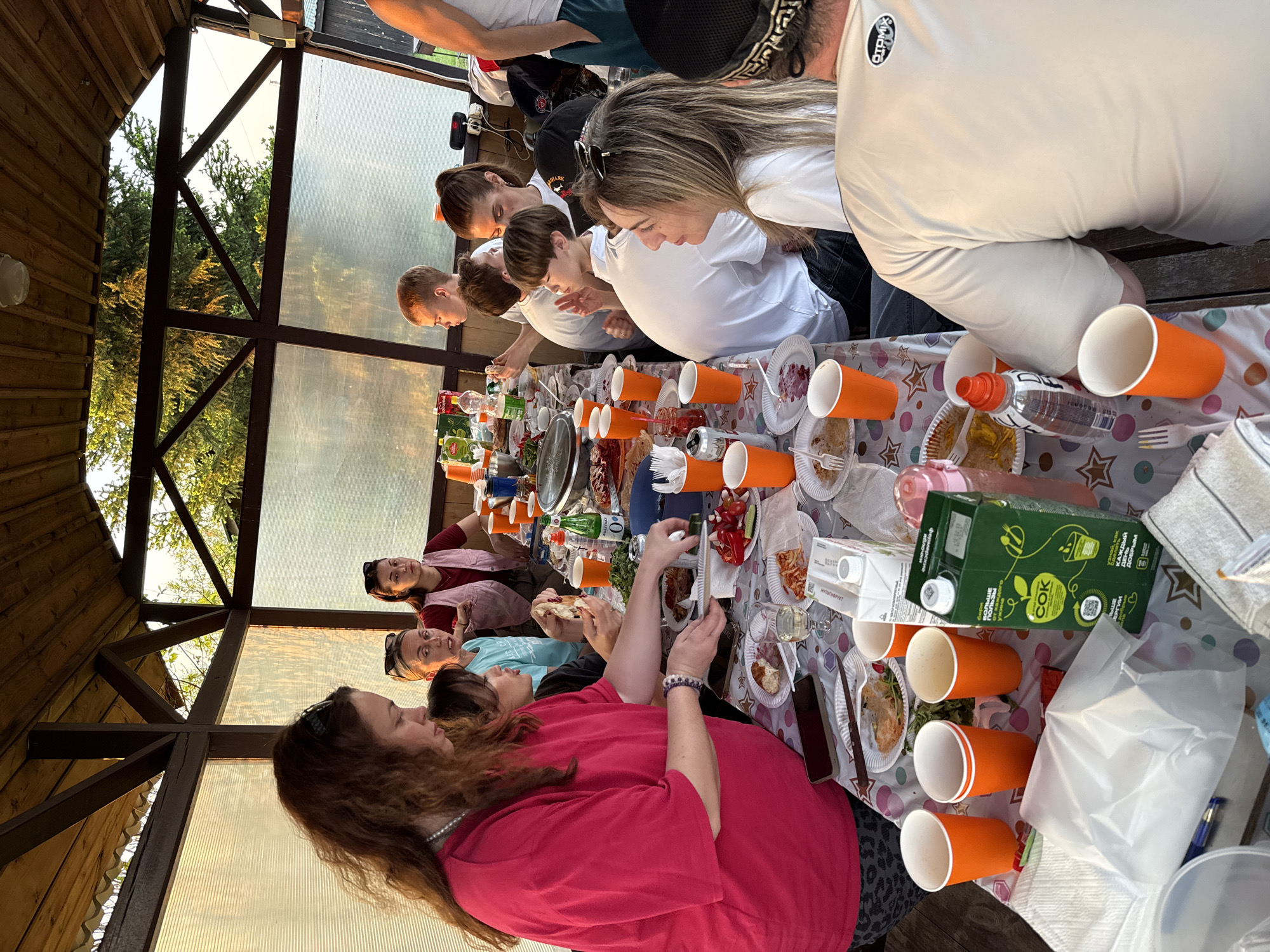 The image shows a group of people gathered around a long table covered with food and drinks in what appears to be an outdoor covered structure, possibly a gazebo or pavilion. The individuals on one side of the table are leaning in, seemingly engaged in eating or looking at the food. The people on the other side of the table are seated and interacting. The setting appears to be a casual gathering with orange cups, bottled water, juice boxes, and plates of food scattered across the table. The structure has wooden supports and transparent walls, revealing trees and foliage outside. The location might be in Кузнецово, Russia, although specific landmarks are not visible in the image to confirm this.