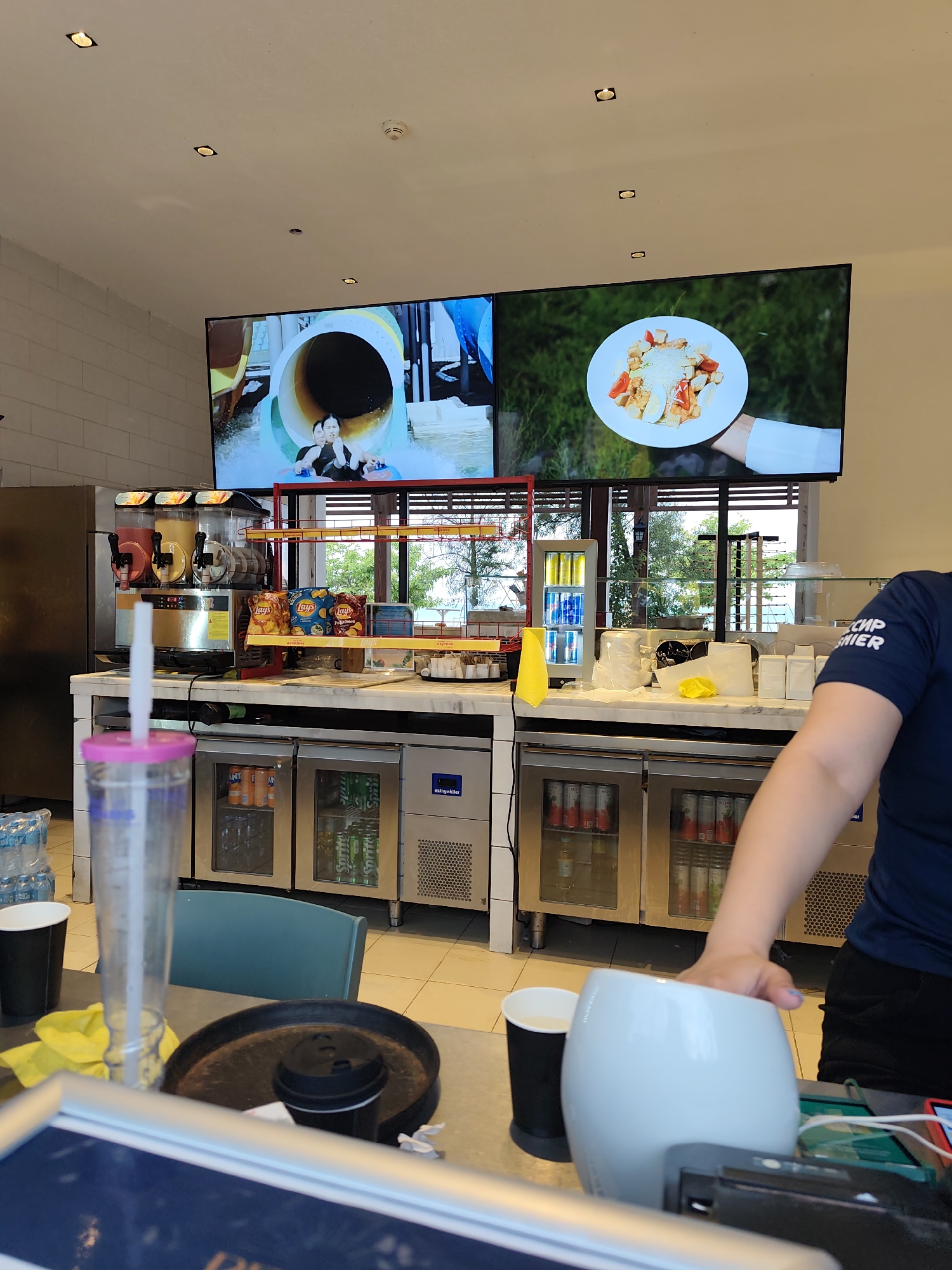 The image depicts an indoor scene that appears to be a food service establishment. In the background, two large screens display content: one shows people on a water slide, and the other displays a plate of food. In the middle ground, a counter area is visible with various food and beverage items, including a slush machine, bags of chips, and beverages in refrigerators. In the foreground, a person's arm and shoulder are visible, wearing a dark blue t-shirt. They are reaching towards a white object on the counter. The overall environment suggests a casual dining or quick-service restaurant setting. There is no indication of the city of Aktau, Kazakhstan in the image itself.