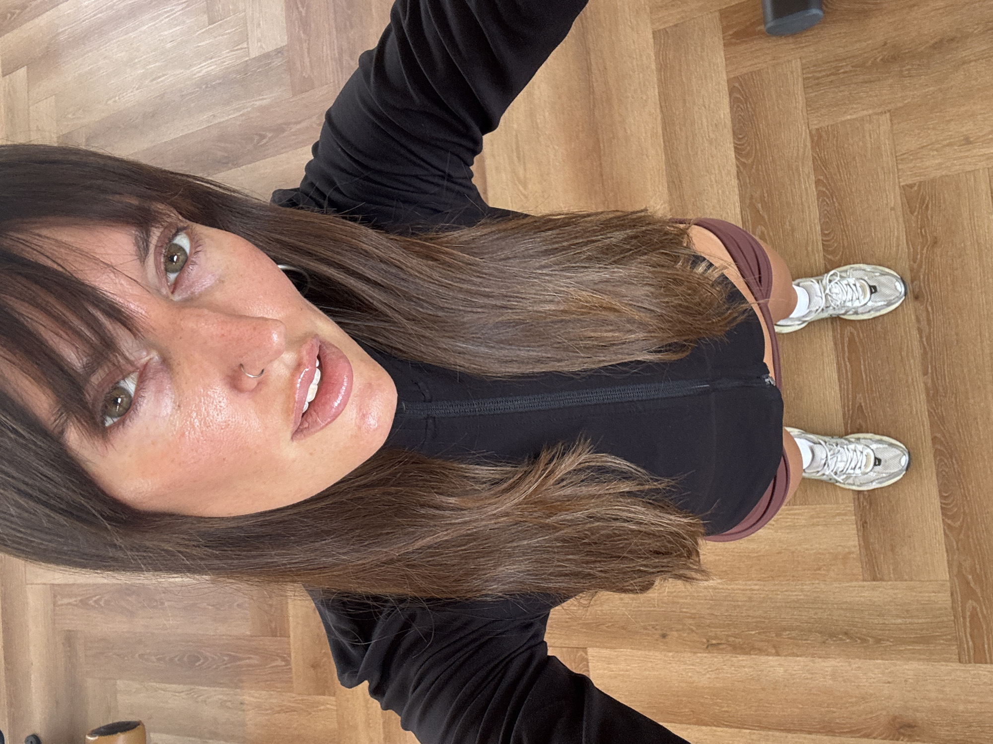A woman, oriented vertically from the viewer's perspective, lies on a hardwood floor. She has long, dark brown hair that falls around her face and shoulders. Her face is turned towards the camera, with one eye visible and her mouth slightly open, revealing white teeth and glossy lips. She wears a black zip-up top, a portion of dark burgundy shorts visible at her waist, and white athletic shoes. The hardwood floor is laid in a parquet pattern. The scene appears to be indoors, possibly a gym or home, given the athletic attire and flooring. The lighting suggests it might be daytime. No visible text is present in the image. The setting is North Kuta, Indonesia.