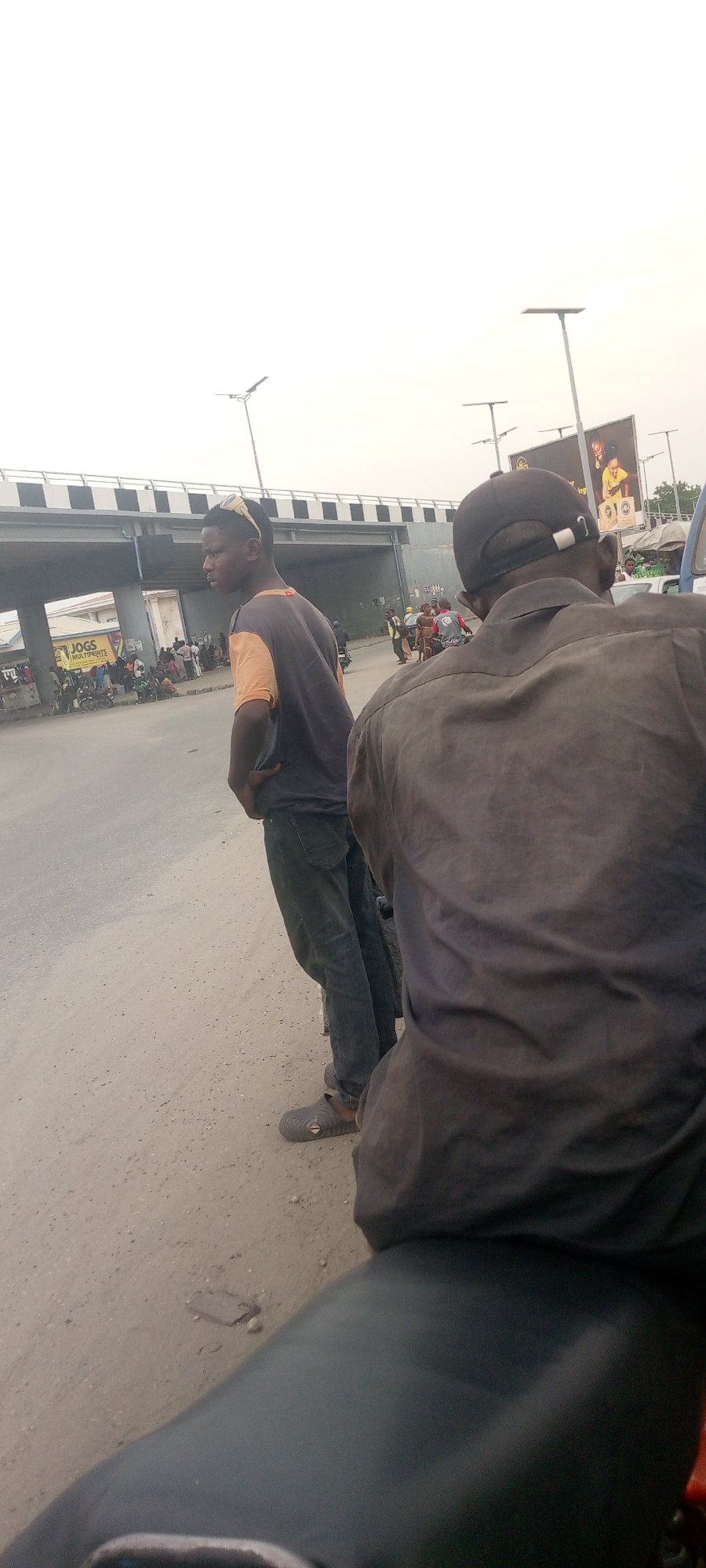 Two men stand on a street in the city of Unknown, Unknown. One man is visible from the waist up and is wearing a dark shirt with an orange shoulder panel, jeans, and shoes. The other man is wearing a dark gray shirt and a baseball cap and is positioned in the foreground. Both men are facing the left side of the image. In the background, there is a bridge over a street, streetlights, and a billboard. People are congregated underneath the bridge. The street surface is unpaved.