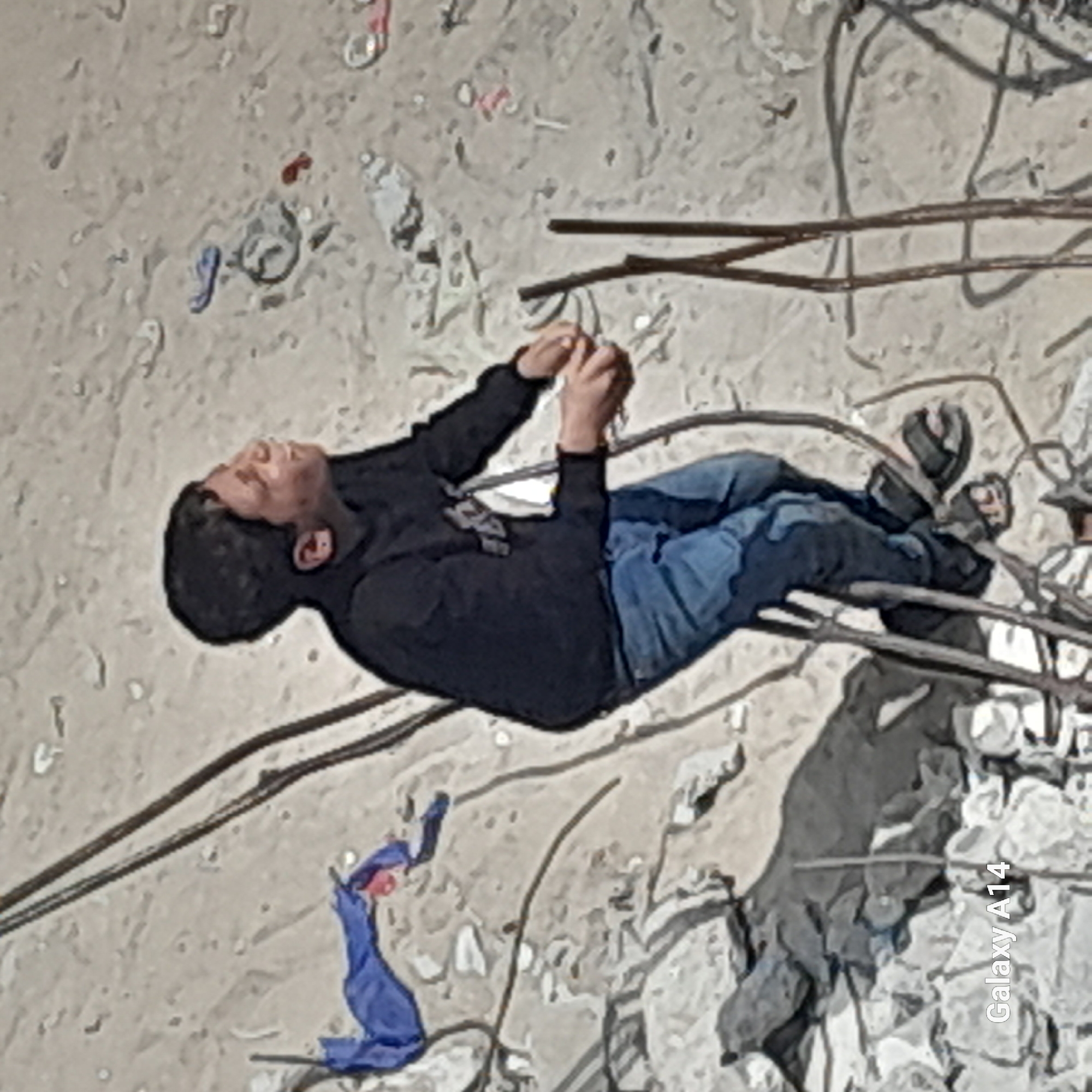 A young male in a black hooded sweatshirt and blue jeans lies on a debris-strewn surface in an unknown city and location. He is surrounded by twisted metal rods and rubble, suggesting a collapsed structure. The boy is holding something in his hands, possibly debris. A scrap of blue fabric is visible near him. A partial caption reading "Galaxy A14" appears in the lower right corner of the image.