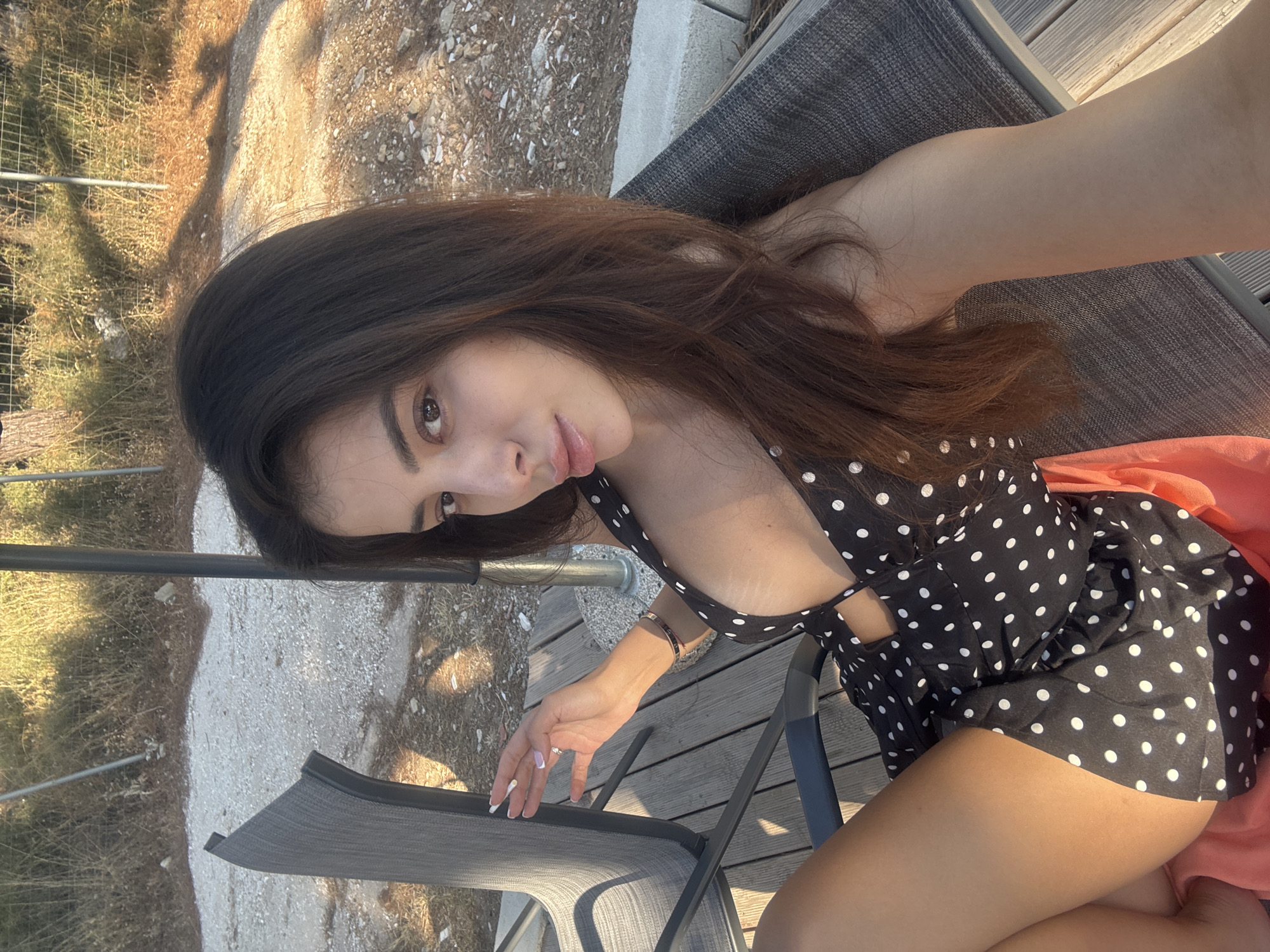 A woman with long brown hair is sitting in a grey outdoor chair on a wooden deck. She is wearing a black and white polka dot dress with a cut-out detail. An orange fabric is draped over her lap. Her right hand is resting on the armrest of the chair, and her left hand is raised, with her fingers extended. The background appears to be a dry, outdoor environment with scattered gravel and vegetation. Sunlight is casting shadows on the ground. The overall impression is a casual, outdoor setting, possibly in a rural or coastal area of Greece.