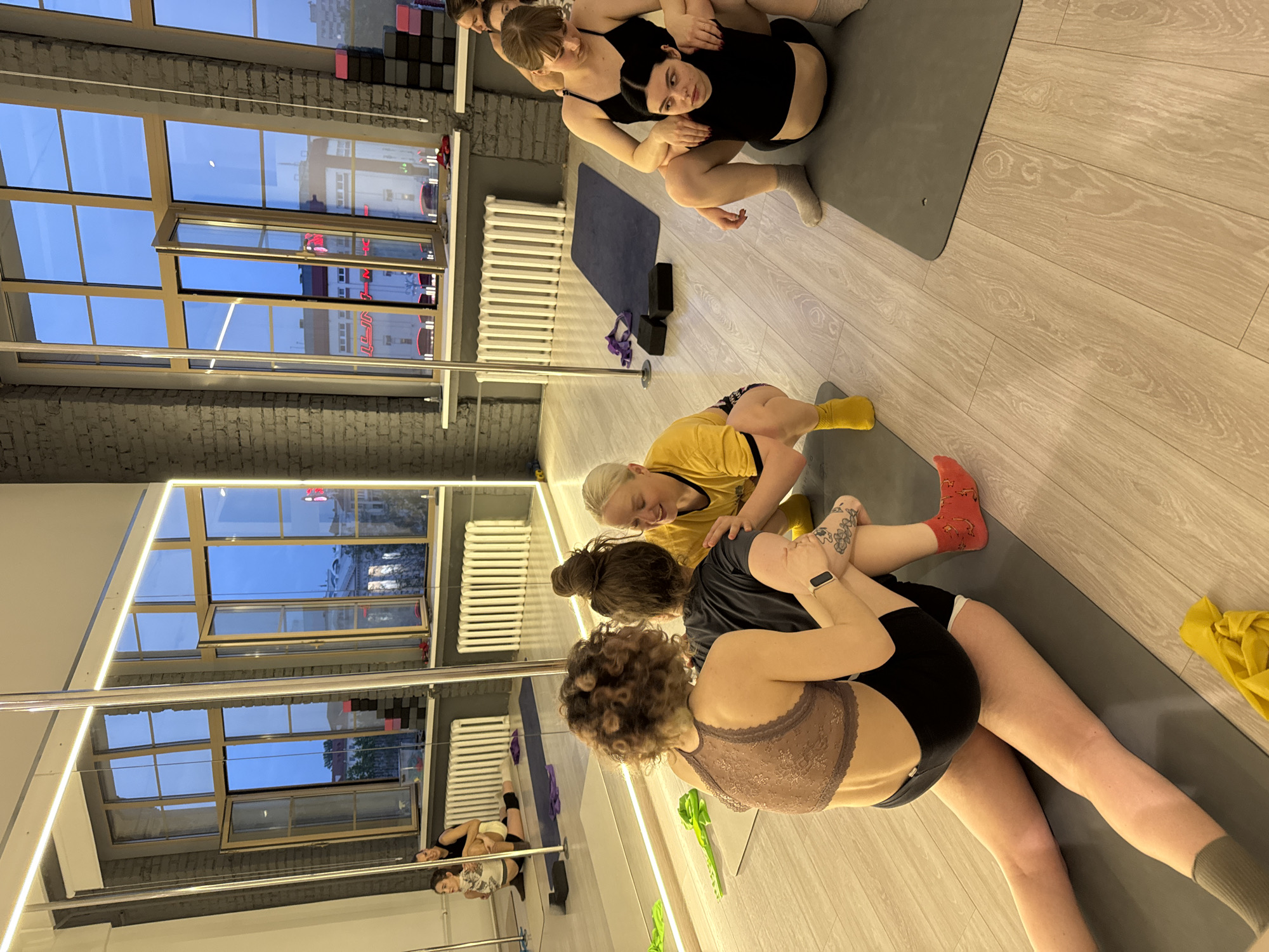 The photo captures a group of women engaged in a stretching exercise within a dance or fitness studio. The studio features a pole dance pole and several windows that suggest it is evening or twilight. Through the windows, the exterior view appears to be a city setting, indicated by buildings and streetlights. Specific landmarks or other defining characteristics are not visible in sufficient detail to pinpoint the exact location.
The location in Minsk, Belarus, is based on available geolocation information. There are no specific landmarks or other defining characteristics visible in sufficient detail to pinpoint the exact location.
One group of women is stretching out while another group is being assisted with stretching. They are using a gray yoga mat and appear to be taking part in some kind of exercise class. The overall setting suggests an urban environment with contemporary architectural elements.