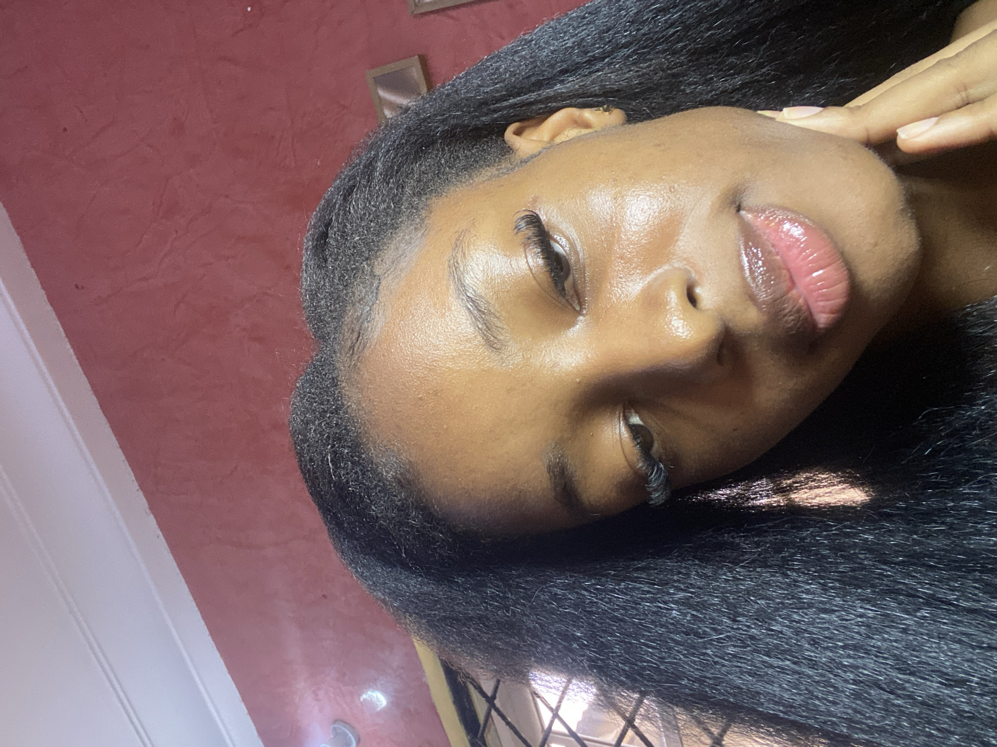 The image is a close-up, low-angle selfie of a young Black woman with long, dark, straight hair. She is wearing subtle makeup, including mascara and lip gloss, and has long, dramatic eyelashes. The woman is touching her chin with her right hand. The background consists of a textured, reddish-pink wall and a white ceiling with molding. There is also a glimpse of a window with a lattice pattern and a framed picture on the wall to the right. The lighting suggests an indoor setting, possibly a home or studio, and casts subtle highlights on the woman's skin, indicating it is well-lit. The image does not provide any specific visual cues that would allow for pinpointing the exact location within Jos, Nigeria.