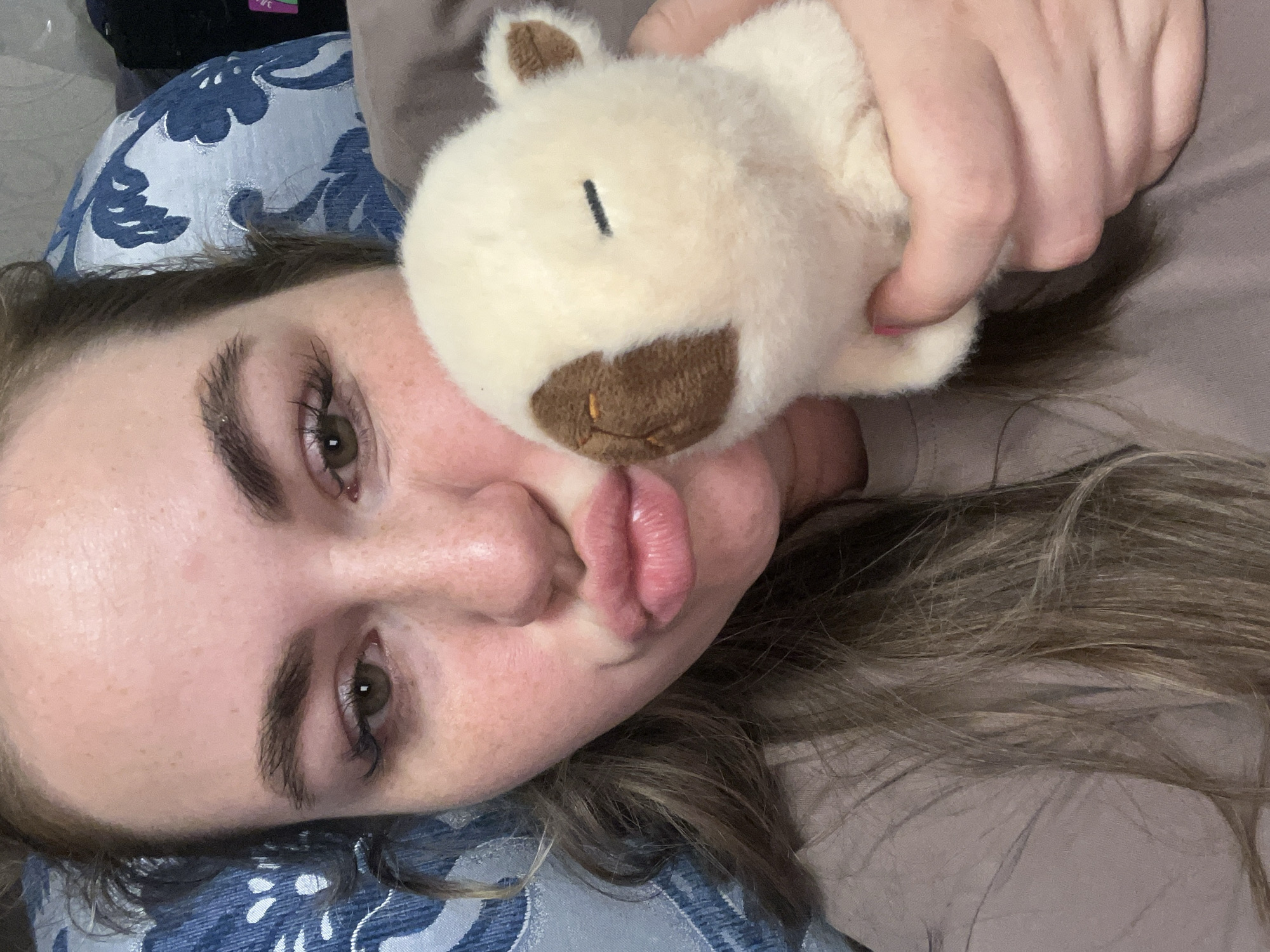 A woman with fair skin, light brown hair, and green eyes is lying down, posing with a stuffed toy bear. The woman is puckering her lips towards the camera. The bear is held close to her face, specifically touching her cheek. The background consists of a blue patterned pillow or cushion on the left side and a beige or light brown surface on the right. The location is not identifiable from the image.