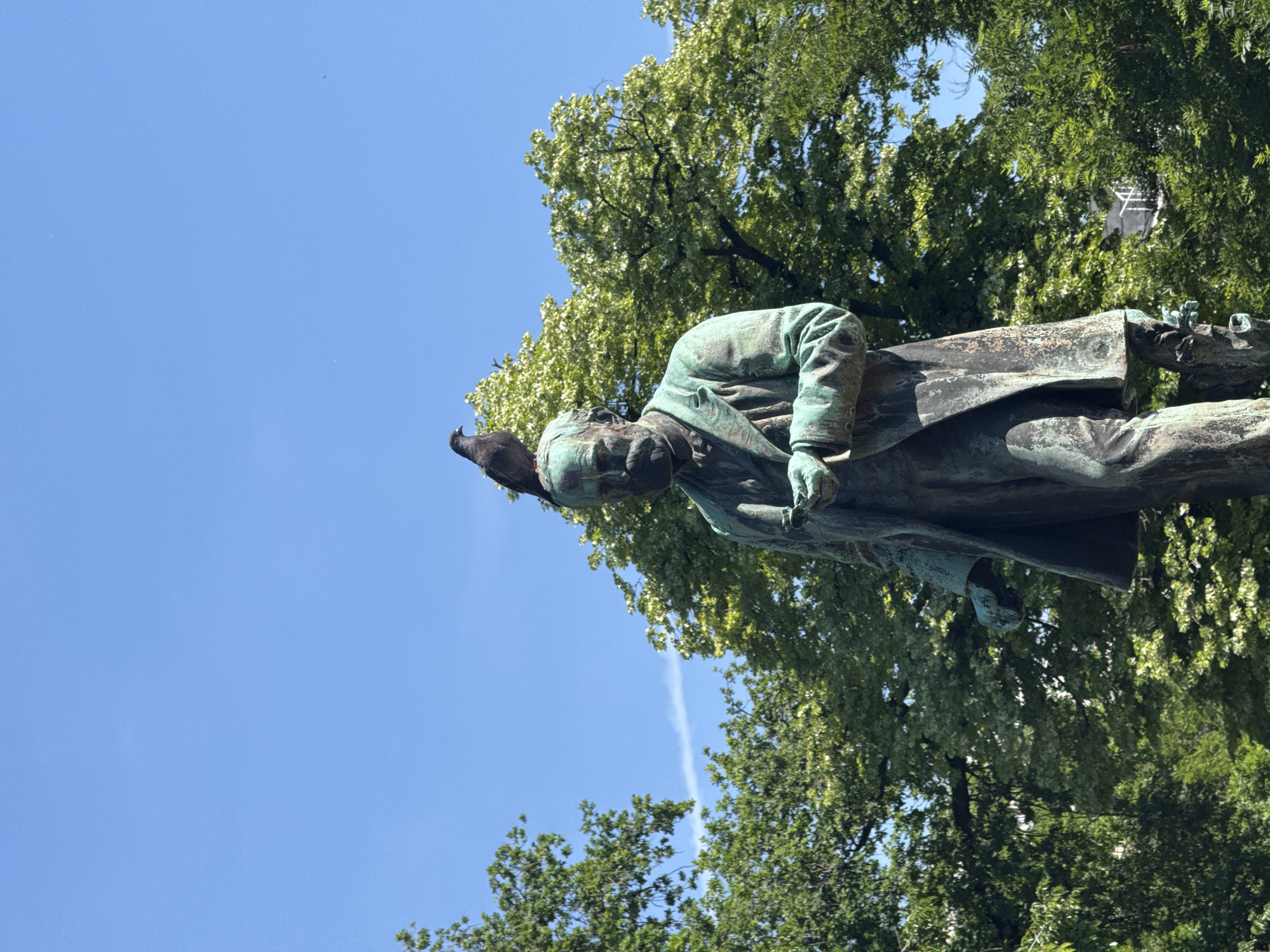 Here is a description of the image:
A statue of a man is pictured outdoors, set against a background of green trees and a blue sky. A pigeon rests on the head of the statue. The statue is weathered and patinated. The statue is located in the city of Belgrade, Serbia.