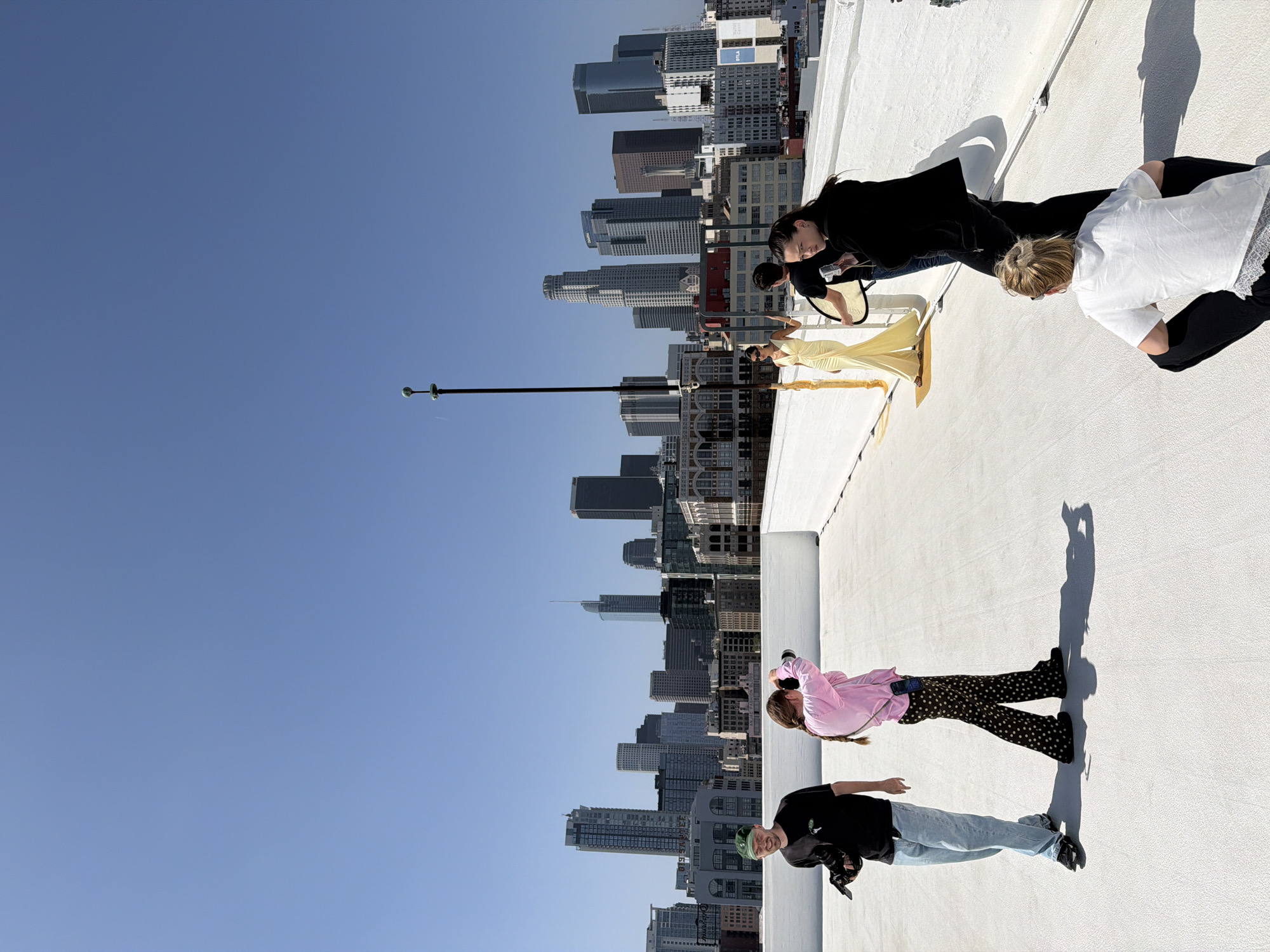 The image captures a rooftop scene with a clear blue sky and a cityscape in the background. The cityscape is identified as Los Angeles, USA, based on the recognizable high-rise buildings. A tall pole extends upwards from the roof, reaching towards the sky. On the rooftop, several individuals are present. One person is wearing a yellow dress and is interacting with another person who is wearing a black top. Another individual is dressed in a pink shirt and spotted pants, while another is wearing a black shirt and light blue jeans. It appears that some of the individuals are taking photographs or are being photographed.