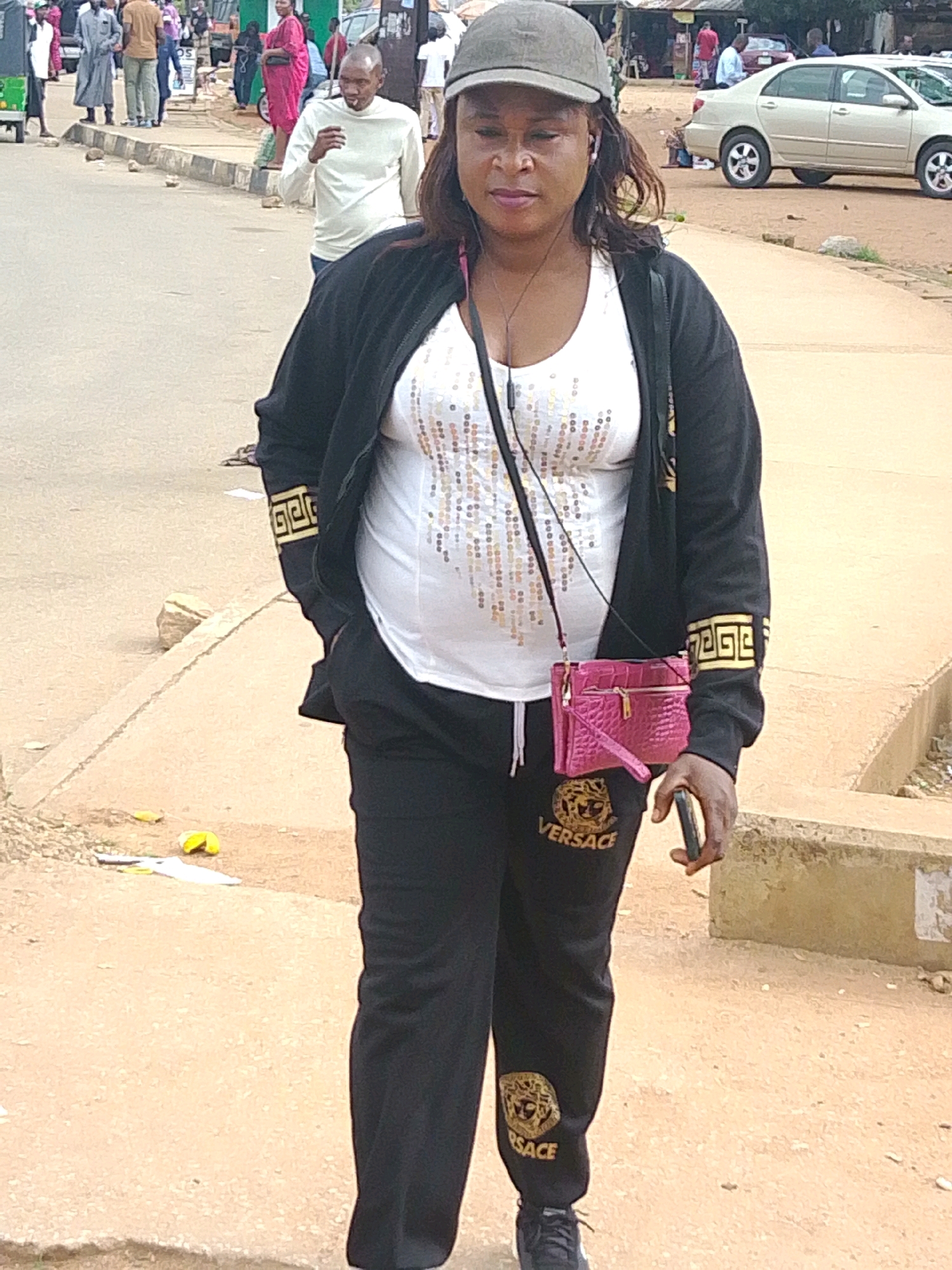 A medium shot shows a woman from the chest down, walking towards the camera on a dirt path in Barakin Lerawi, Nigeria. She wears a black hooded jacket with gold Greek key patterns on the sleeves, a white tank top adorned with sequins, and black tracksuit bottoms with gold Versace logos. A pink crocodile-textured purse is slung across her body, and she holds a dark smartphone in her right hand. Her left hand is in the pocket of her jacket. Behind her, a street scene unfolds with other pedestrians, vehicles, and shops. The ground is paved with a lighter colored concrete edge along the dirt path. In the background, a beige sedan is parked on the street.
