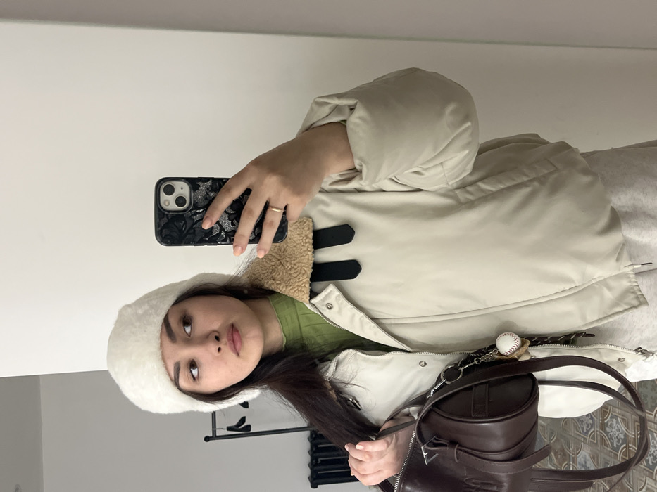 This image is a mirror selfie taken indoors in Neftekamsk, Russia. It features a female subject, depicted from the mid-torso upwards, wearing a white faux-fur hat, a light green ribbed turtleneck, a cream-colored puffer jacket with a tan faux-shearling collar and black strap accents, and light grey sweatpants. She holds a dark-colored smartphone with an elaborate, possibly floral or animal-patterned case in her right hand, revealing a gold-colored ring on her right ring finger. Her left hand holds a dark brown, barrel-shaped handbag with a silver-toned emblem and a keychain adorned with a miniature baseball and other charms. The subject gazes off to her right. The environment, reflected in the mirror, consists of white walls, a dark garment rack with a hanger on the left, a dark radiator below it, and a patterned tiled floor.