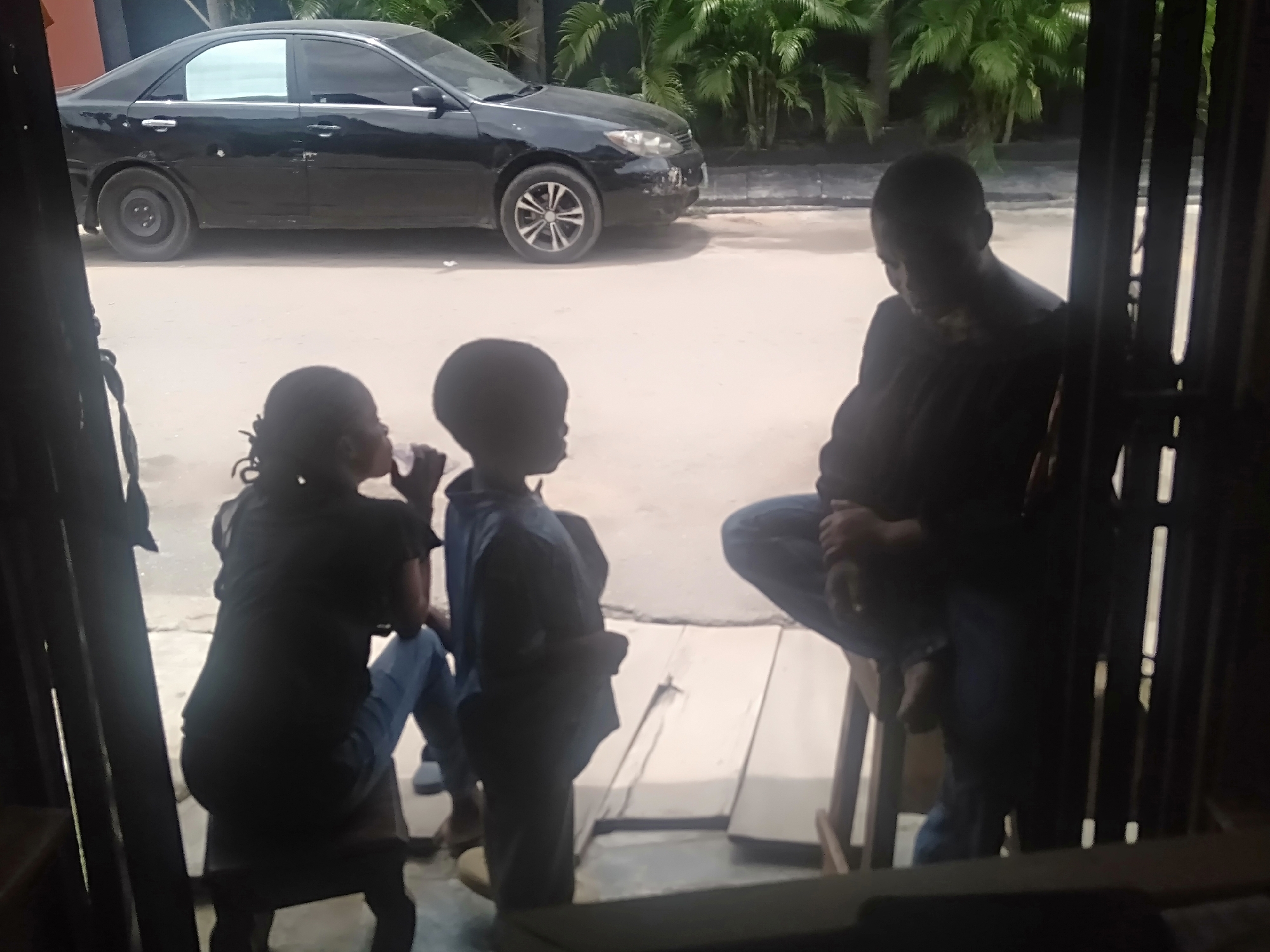 The image depicts a group of people standing outside, partially obscured by a building structure. A black car is parked on the road in front of the group, with green foliage visible in the background. One individual is seated on a stool. The specific location within the city of Unknown, Unknown is not discernable from the image.