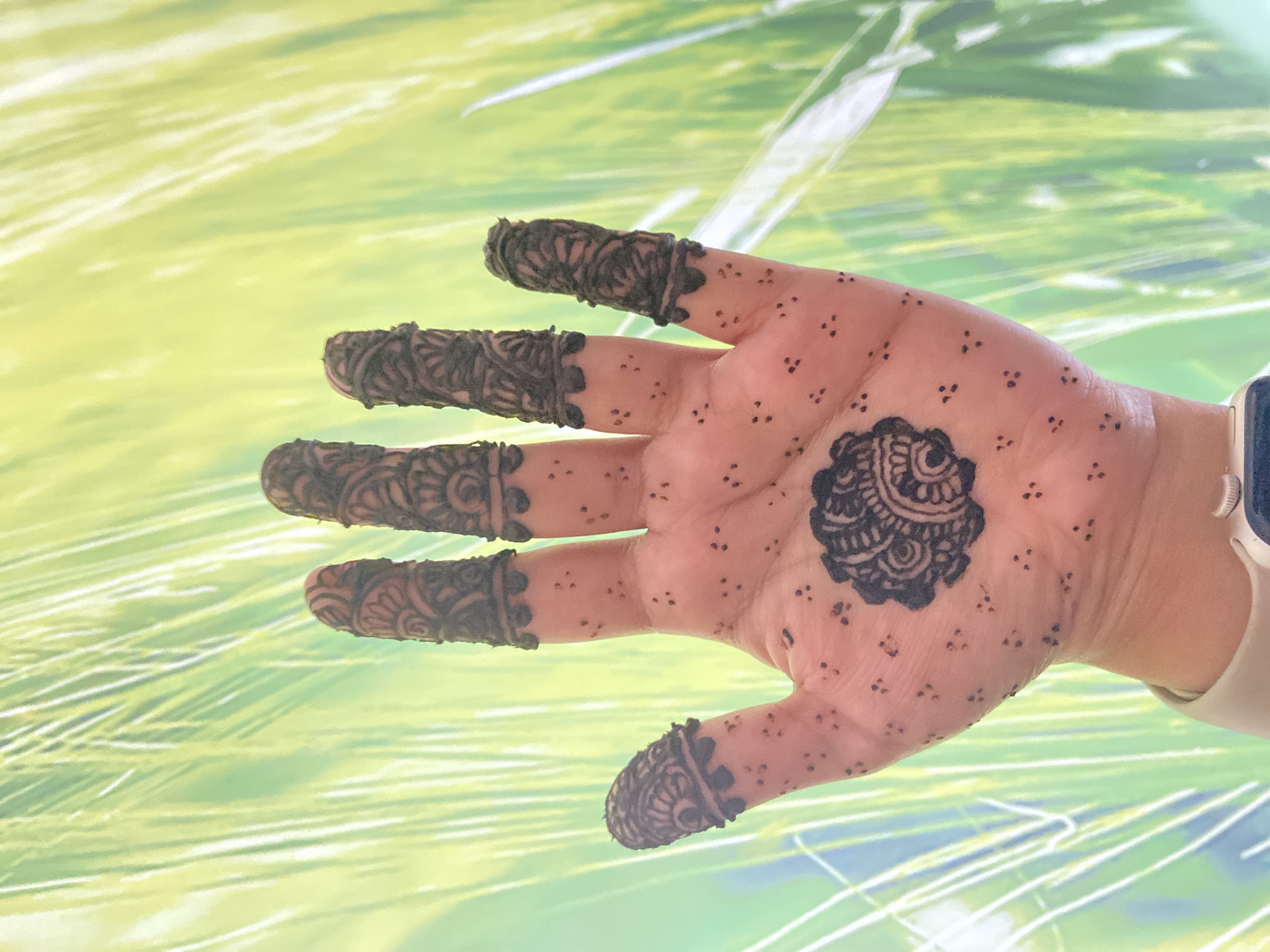 A hand with dark henna designs is positioned in front of a blurred background of green and yellow. The henna covers the fingertips of each finger and includes a larger circular design on the palm. A watch is visible on the wrist. No specific location in Dhaka, Bangladesh can be determined from the image.