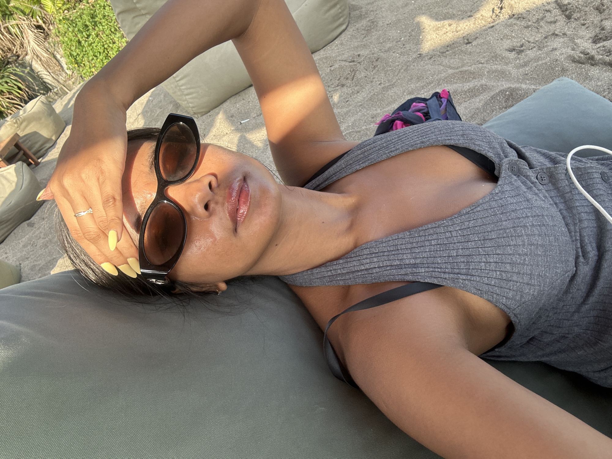 A woman is lying on a lounge chair on a sandy beach in North Kuta, Indonesia. She is wearing a grey ribbed tank top and sunglasses. Her hand is raised to her forehead, shielding her eyes from the sun. Her fingernails are painted a bright yellow. The sun is casting a shadow across her face and arm. The background shows more lounge chairs, sand, and some greenery.