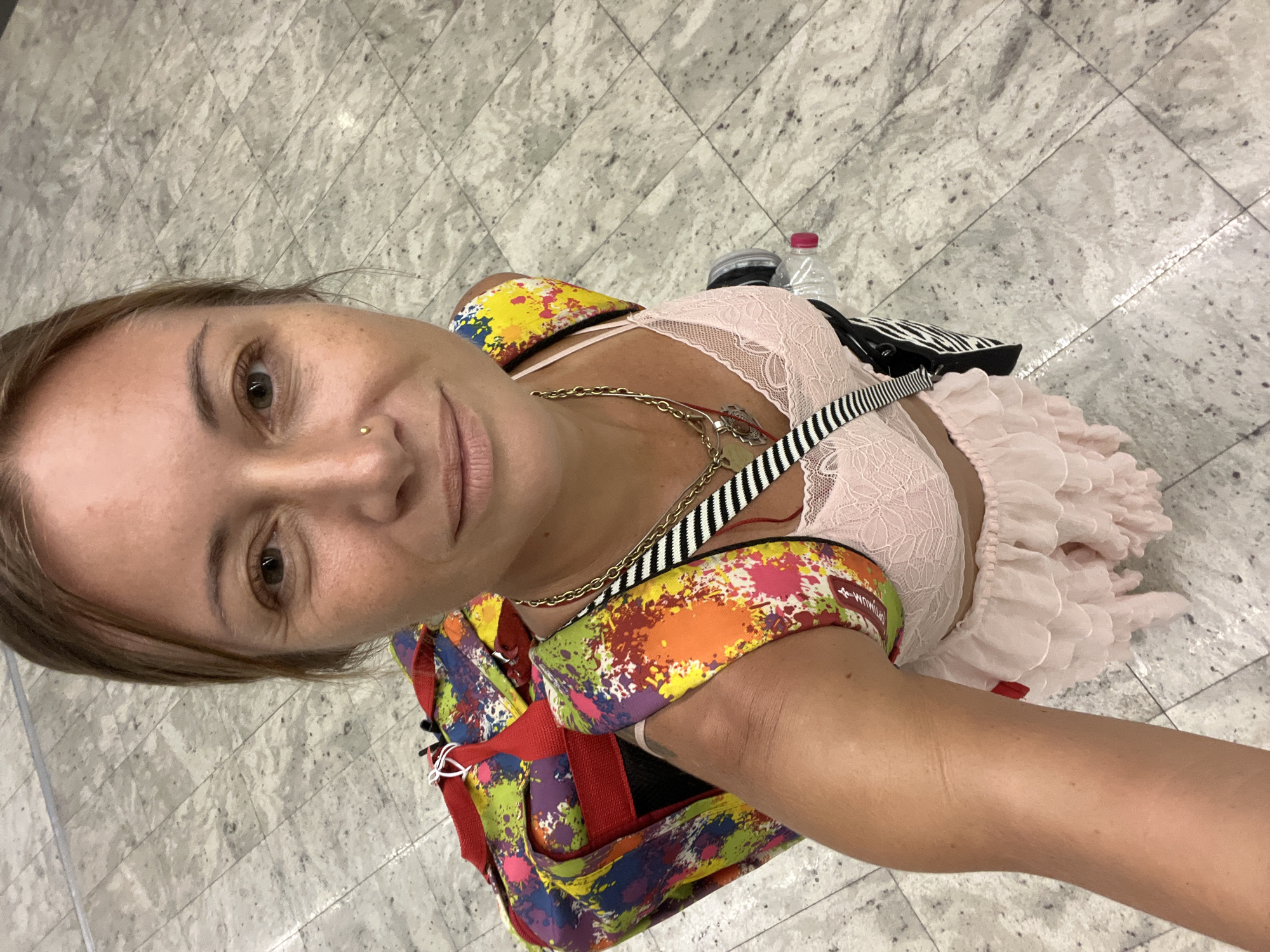 In Katunayake, Sri Lanka, a woman is photographed from a high angle, looking up at the camera. She has light brown hair pulled back, a small nose ring, and is wearing a pink lace bralette and a light pink ruffled skirt. A colorful, paint-splattered duffel bag is slung over her right shoulder, with one strap visible on her left. A gold chain necklace with a pendant and a black and white striped strap from another bag are also visible around her neck. She is standing on a light gray tiled floor with a diamond pattern. A clear plastic water bottle with a pink cap lies on the floor near her right leg.