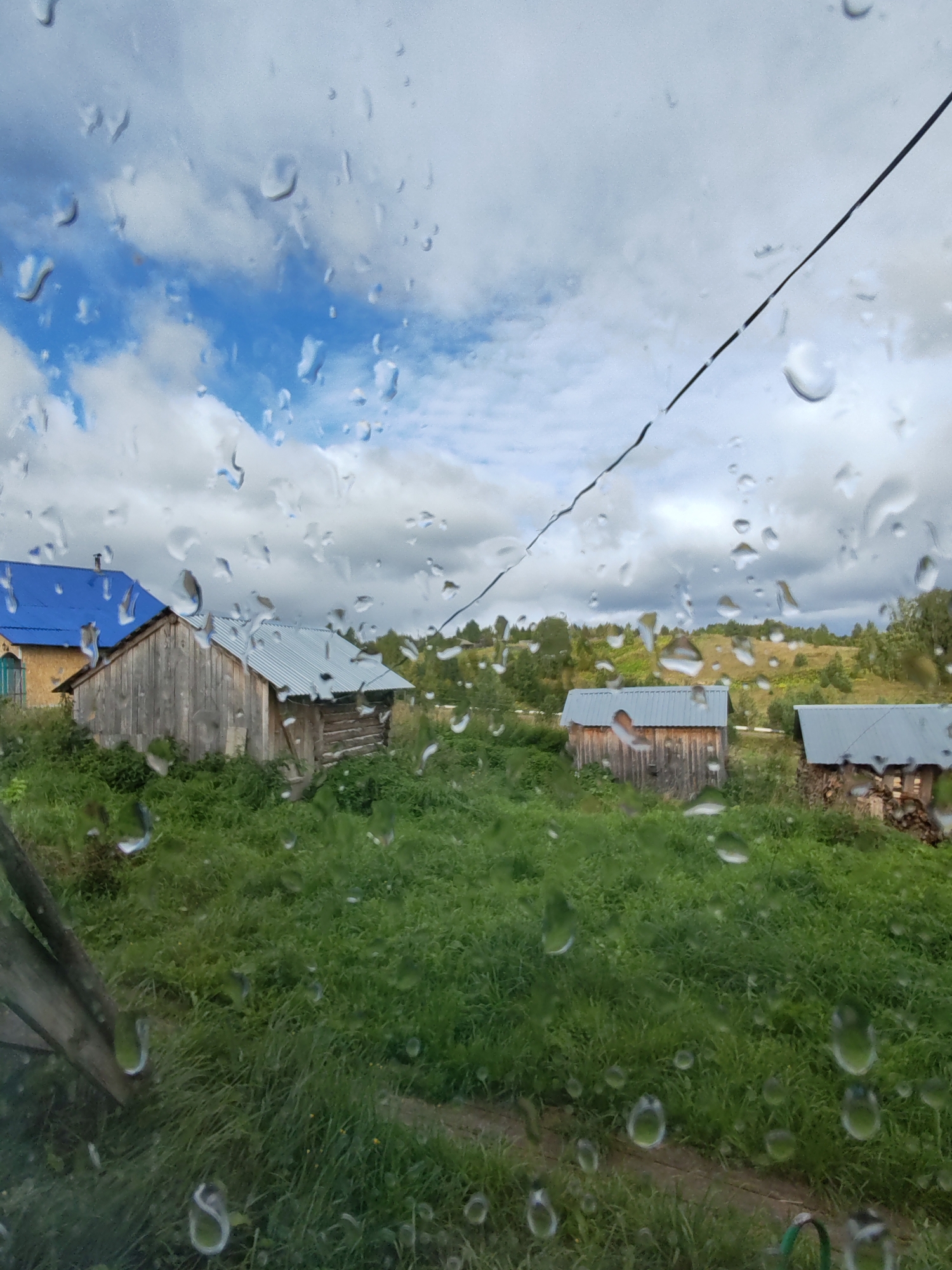 This is an outdoor, eye-level, medium shot of a village scene in Chuchlom, Russia, taken through a window with raindrops on the glass. The foreground is dominated by the distorted view of a lush green meadow and a dirt path. In the middle ground, several wooden structures with corrugated metal roofs are visible. To the left, a house with a bright blue roof stands partially obscured by a rustic wooden fence post. Behind the house, there is a dense area of green vegetation and trees. Further in the distance, rolling hills covered in green foliage and scattered trees stretch across the landscape under a cloudy sky with patches of blue. A dark wire runs diagonally across the upper portion of the image. The presence of raindrops on the window creates a blurred and fragmented perspective of the entire scene. There are no people or animals visible in the image.