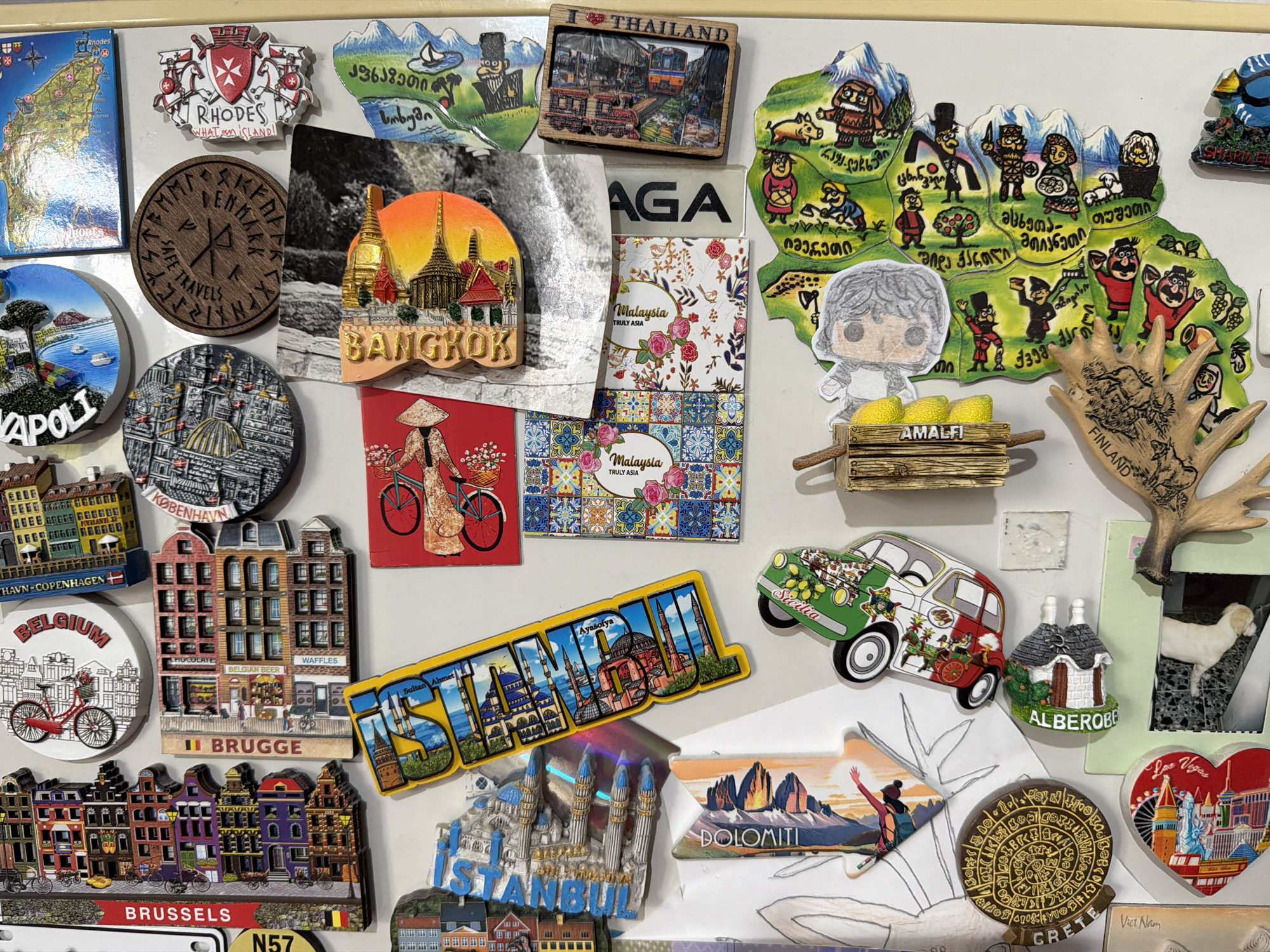 This close-up, top-down shot captures a densely packed collection of colorful souvenir magnets displayed on a white surface, likely a refrigerator door. The scene suggests a home in Tbilisi, Georgia, as indicated by a prominent map-shaped magnet featuring cartoon figures and Georgian script.
The magnets are diverse in shape, size, and material, showcasing numerous travel destinations and cultural icons. Visible magnets include: "Rhodes" (a map and a coat of arms), "NAPOLI," a wooden "SAFE TRAVELS" magnet with runes, "THAILAND" (a framed train scene), "BANGKOK" (golden temples), and "Malaysia TRULY ASIA" (floral designs).
European locations are well represented with "AMALFI" (lemons in a crate), "FINLAND" (an antler), "KØBENHAVN" (Copenhagen buildings), and "BELGIUM" with magnets for "BRUGGE" and "BRUSSELS" depicting characteristic architecture, bicycles, and references to Belgian beer and waffles. Further magnets include "ISTANBUL" (a word magnet featuring Sultan Ahmet and Ayasofya, and a 3D mosque), "Sicilia" (a car magnet), "DOLOMITI" (mountains with a skier), "ALBEROBELLO" (a trullo house), "CRETE" (a replica of the Phaistos Disc), and "Las Vegas" (a heart-shaped design). A partially visible "Viet Nam" magnet is also present, along with a "AGA" magnet. A hand-drawn Funko Pop-style character is affixed over part of the Georgia map. The collection signifies extensive travel and cherished memories from around the world.