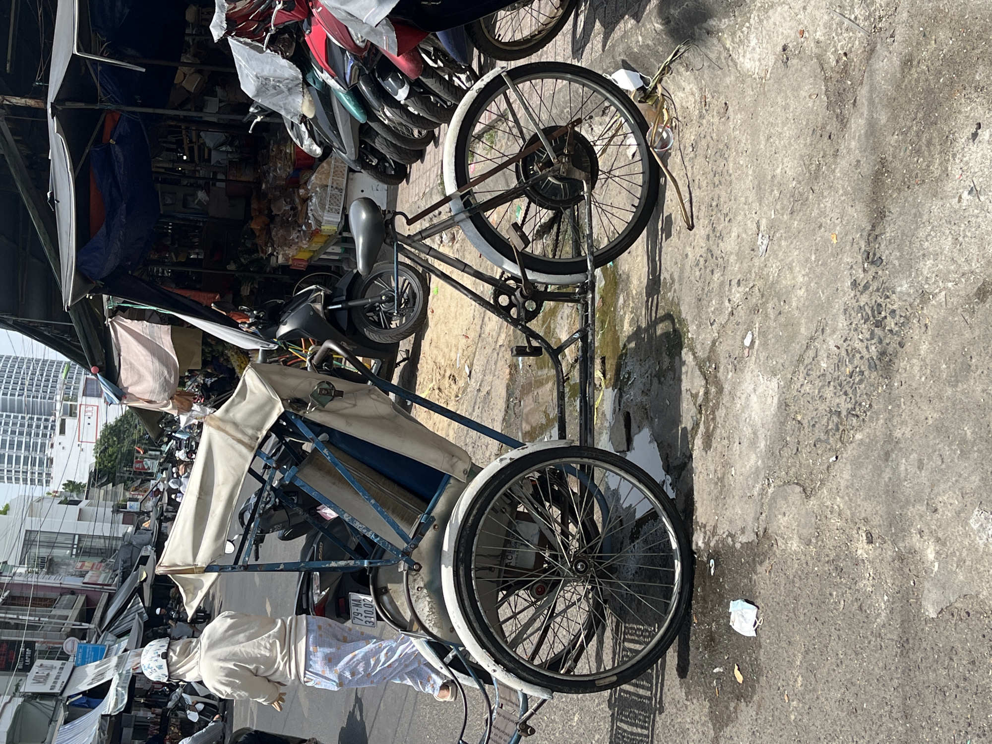 The image captures a street scene in Nha Trang Ward, Vietnam, likely a marketplace or busy thoroughfare. In the foreground, a three-wheeled cycle rickshaw is parked on a wet, paved surface littered with debris, including a discarded face mask. The rickshaw has a blue frame and a white fabric canopy covering its passenger area.
To the left of the rickshaw, a person wearing a light-colored hooded jacket, light-colored patterned pants, and a white helmet is walking away from the camera. Behind the rickshaw and the person, a motorcycle with a license plate reading "79-NA 310.02" is visible.
The background reveals a bustling urban environment with market stalls and shops. Various motorcycles are parked in rows in front of these stalls. The stalls are covered with awnings and tarps, displaying goods for sale. Buildings line the street, and a tall modern building is visible in the distance. The overall atmosphere suggests a hot, sunny day.