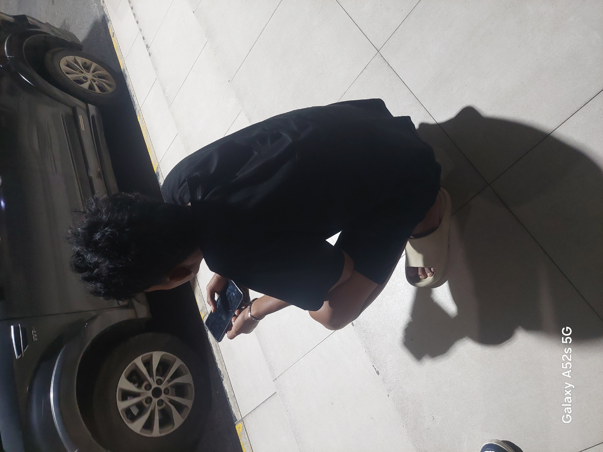 In an outdoor scene in Bumisari, Indonesia, a person is crouching on a tiled surface. The individual is wearing a black t-shirt and shorts, and their back is to the camera. They are holding a smartphone in their hands, appearing to be using it. A dark-colored vehicle is parked behind them, with its rear tire visible. The ground is covered in light-colored tiles, with a yellow and black striped border running along the edge of the road. Shadows are cast on the tiled surface, indicating a light source from above. The overall lighting suggests it is either dusk or nighttime, with artificial lights illuminating the area. A watermark at the bottom of the image reads "Galaxy A52s 5G".