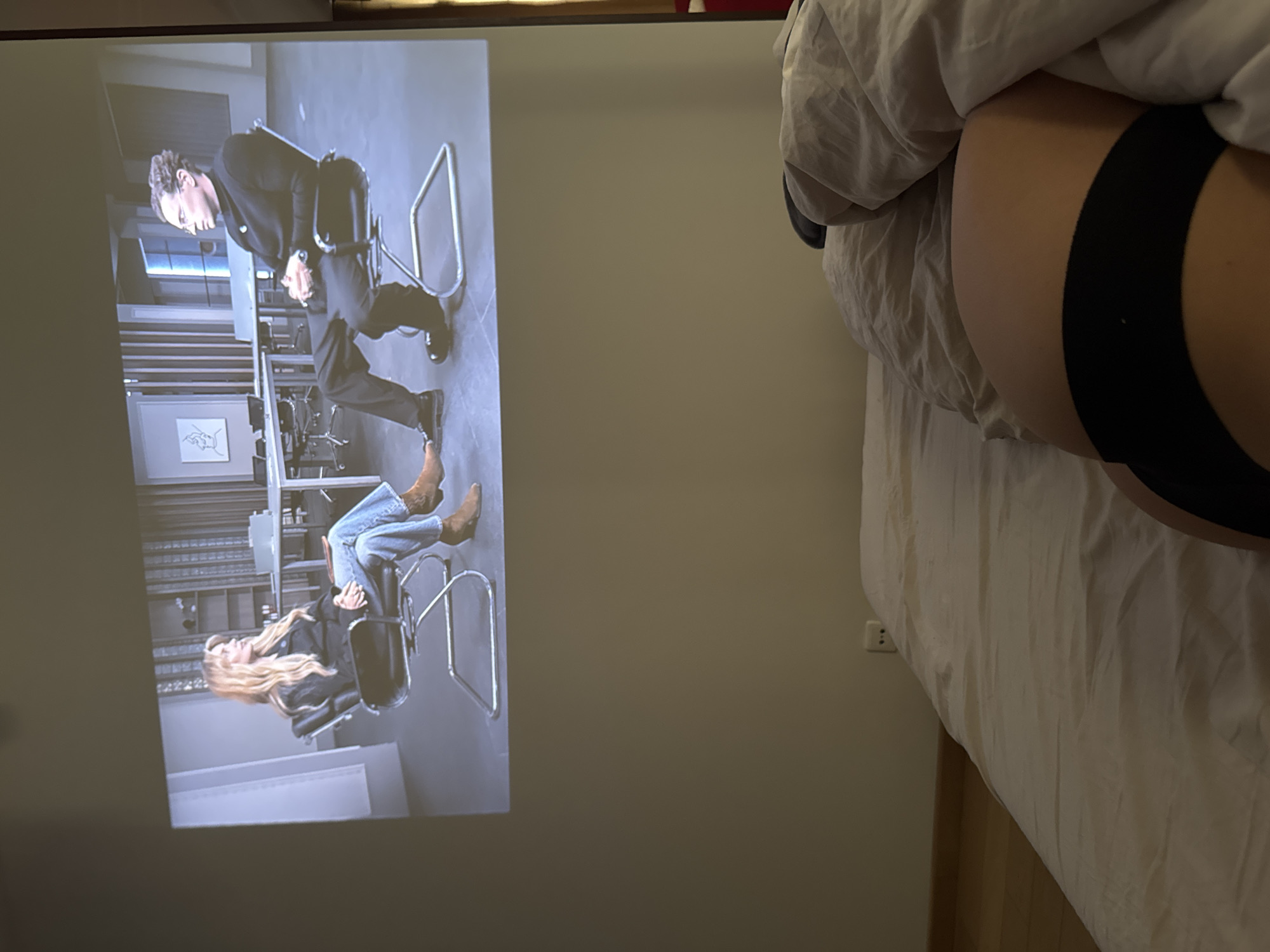 The image shows a partially obscured view of a bed with a person lying in it, partially covered by bedding. The person is wearing black underwear. On the wall, a projector displays a scene showing a man in a suit standing over a woman sitting in a chair, seemingly in an office setting. The location is likely a private residence, but the city of Warsaw, Poland, cannot be confirmed from the provided image.