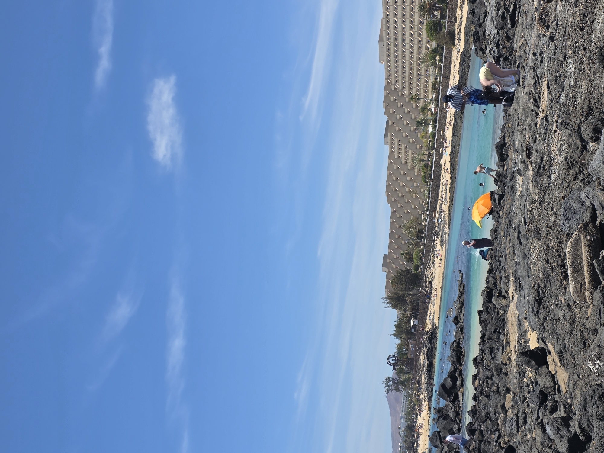 This is a wide-angle, eye-level shot of a rocky coastline with a turquoise ocean and a beach in the background. The scene is bathed in bright sunlight, suggesting a clear, warm day. The sky is a vivid blue with a few wispy white clouds scattered across it.
In the foreground, dark, volcanic rocks create a rugged texture along the edge of the water. Several people are visible on these rocks, some sitting or bending over, appearing to be getting ready for or recovering from a swim. One person, wearing a striped shirt and patterned shorts, stands near the water's edge. An orange umbrella is set up on the rocks, providing a splash of color against the dark stone and blue water.
The turquoise water is clear and inviting, with gentle waves lapping at the shore. Further back, a sandy beach is lined with palm trees and sunbathers. Beyond the beach, a large, multi-story hotel or resort complex with a distinctive facade dominates the background. Further to the left, a windmill and more buildings are visible, along with a distant mountain range.
The overall atmosphere is one of a relaxed, sunny day at a popular coastal resort. The people seem to be enjoying leisure activities, likely swimming, sunbathing, or simply relaxing by the sea. The location appears to be a well-developed tourist area, with a combination of natural beauty and man-made structures. No visible text is present in the image.